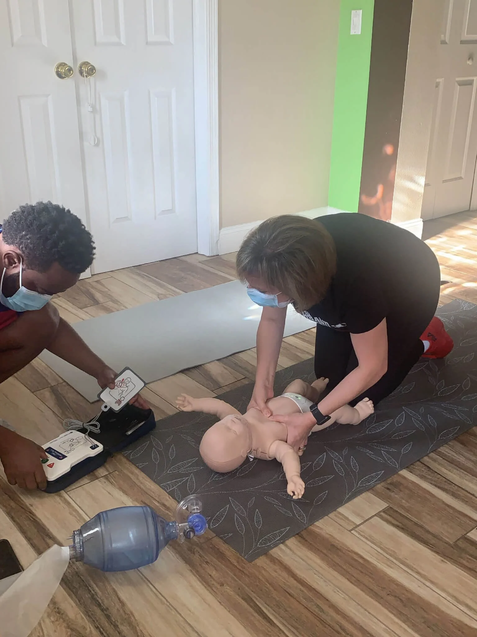 Two instructors practicing infant CPR on a doll, with one using a resuscitation mask and the other performing chest compressions, on a yoga mat in a room with wood floors.