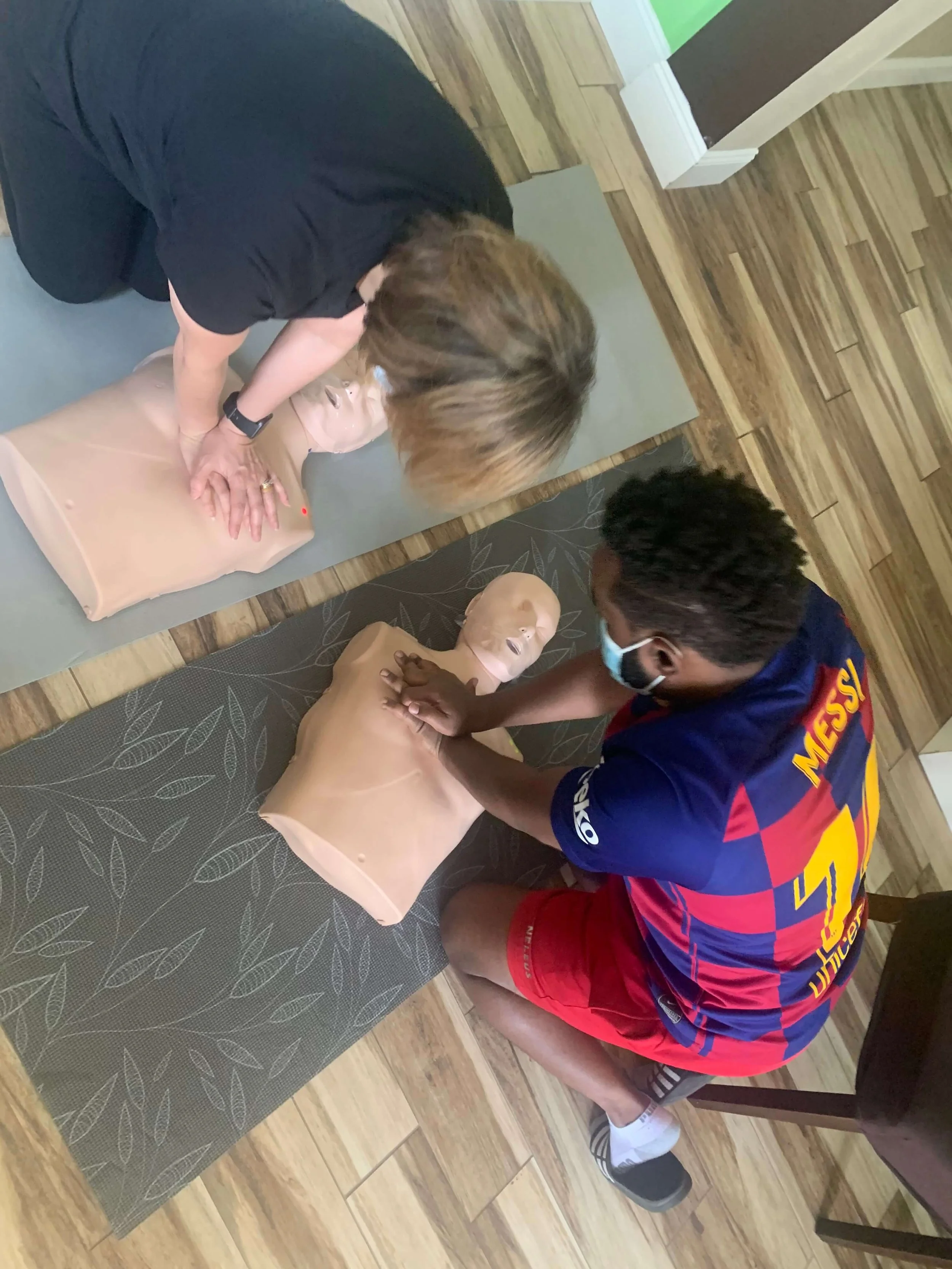 Two people practicing CPR on adult training mannequins in a room with wooden floors, one person kneeling on a gray mat and the other on a patterned dark mat, both performing chest compressions.