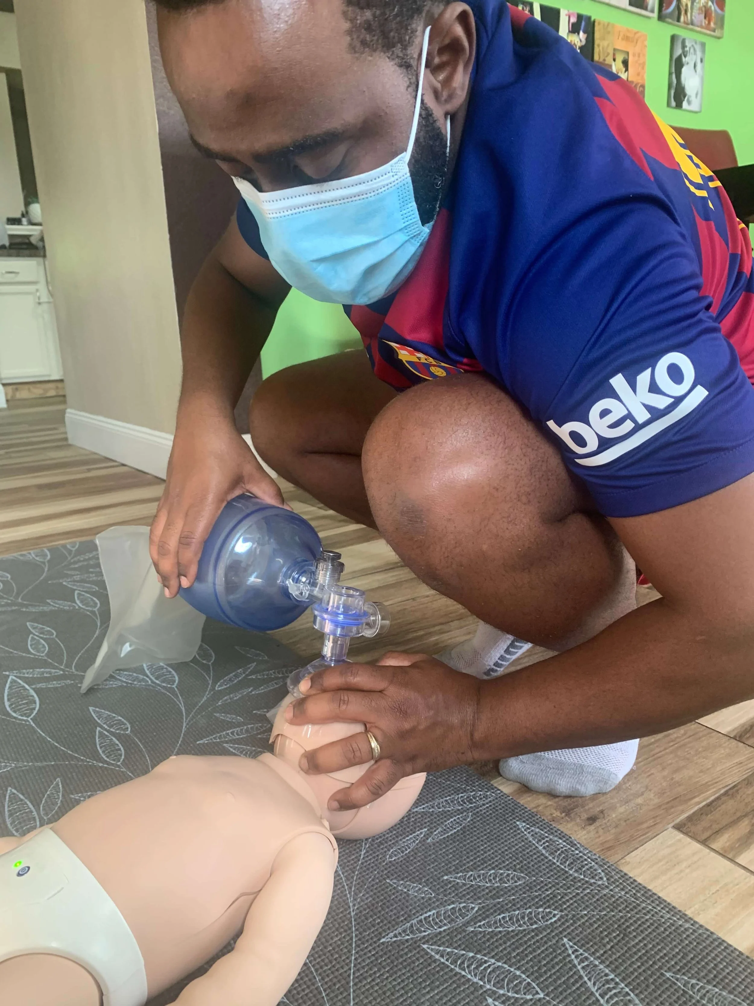 A man wearing a face mask and a blue and red sports shirt is practicing CPR on a mannequin, using a ventilation bag to give air.