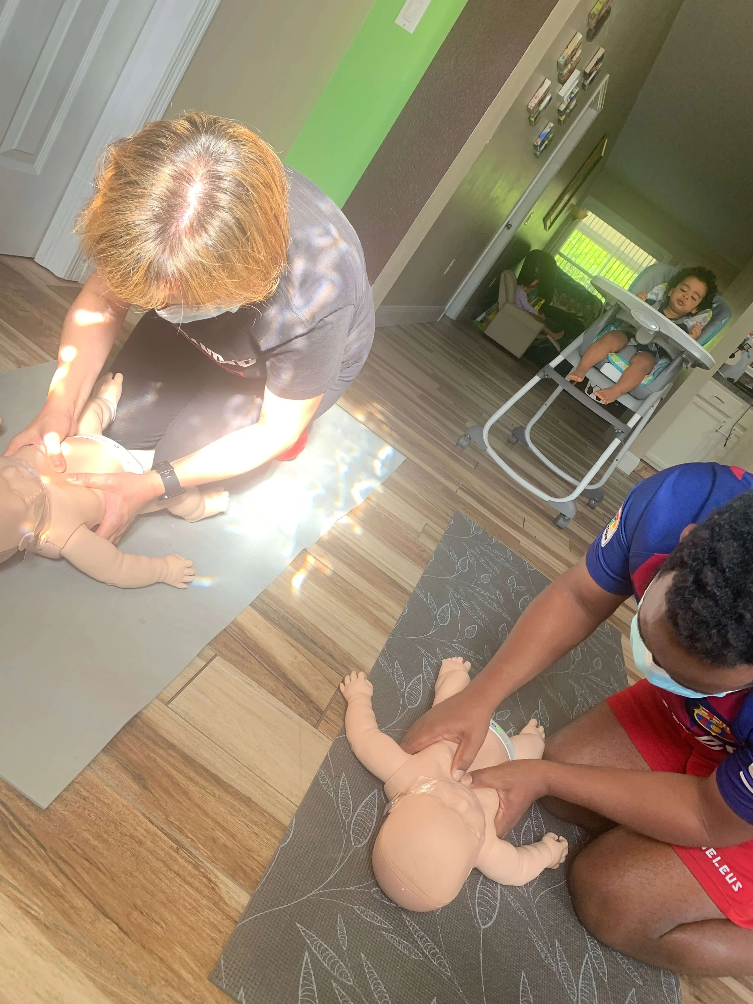 Two people practicing infant CPR on dolls during a training session inside a house, with a child in a highchair and a cozy living space in the background.