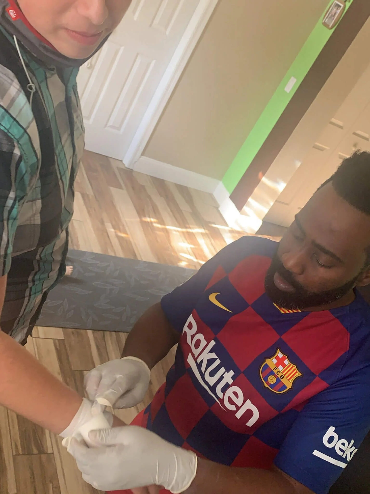 A man in a blue and red Barcelona football jersey is getting a vaccination by a healthcare professional wearing white gloves, in a room with wood flooring and a yoga mat on the floor.