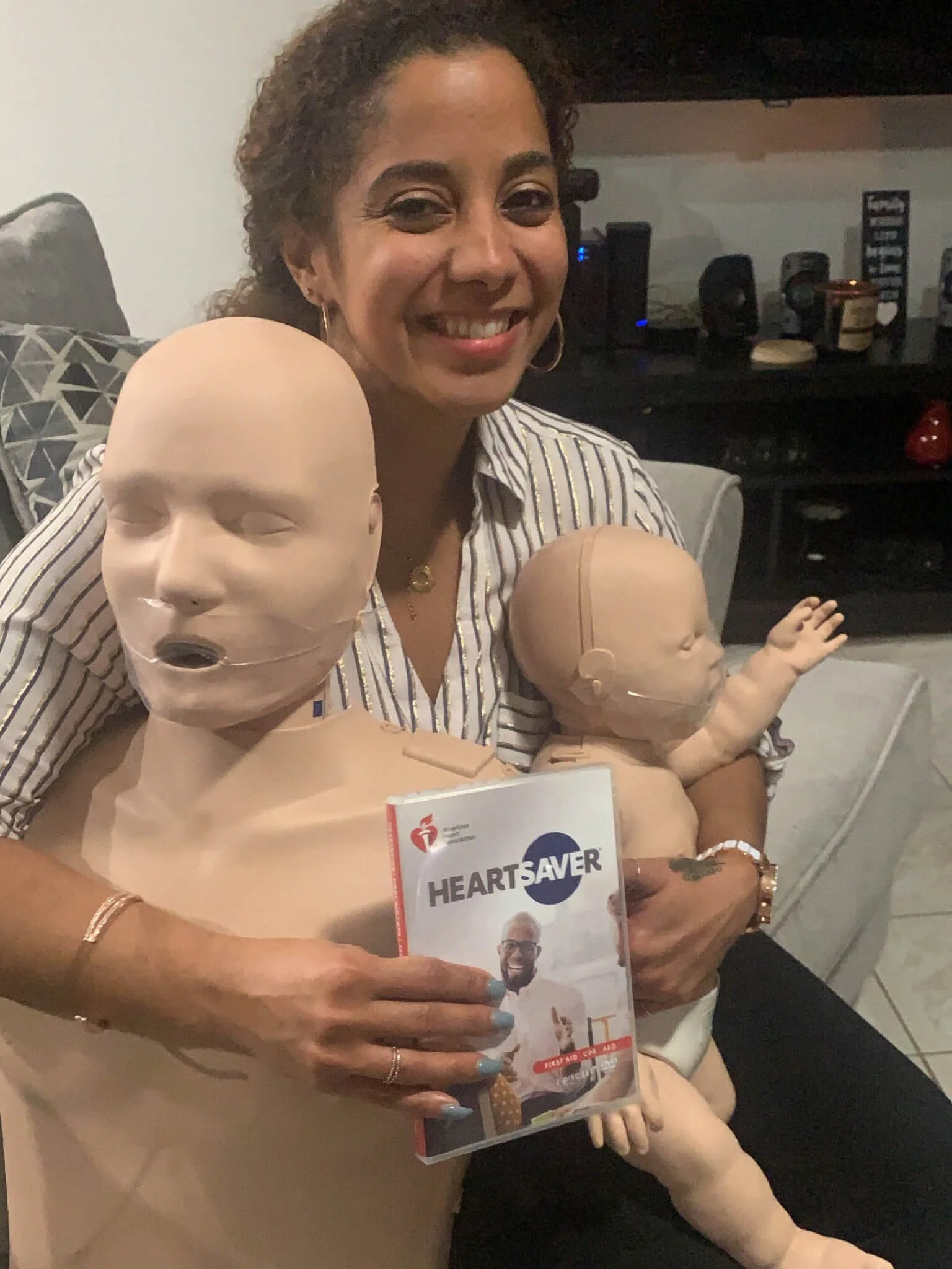 A smiling woman with curly hair holding a medical mannequin and a pamphlet titled 'HeartSAVER'. She is sitting on a couch with another mannequin in her lap. The background shows a television and some electronic devices.