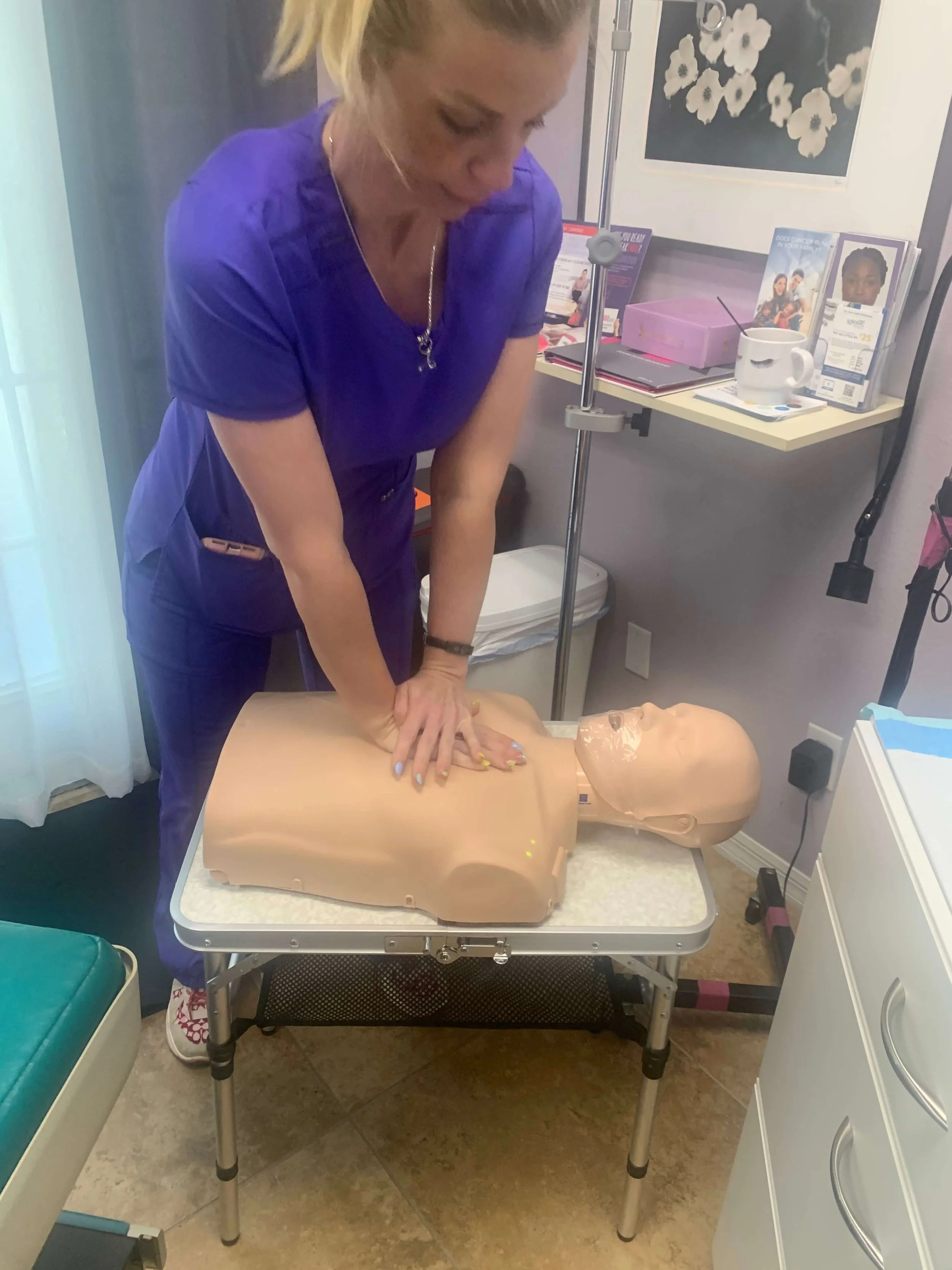 A woman in medical scrubs demonstrating CPR on a medical training mannequin in a clinical setting.