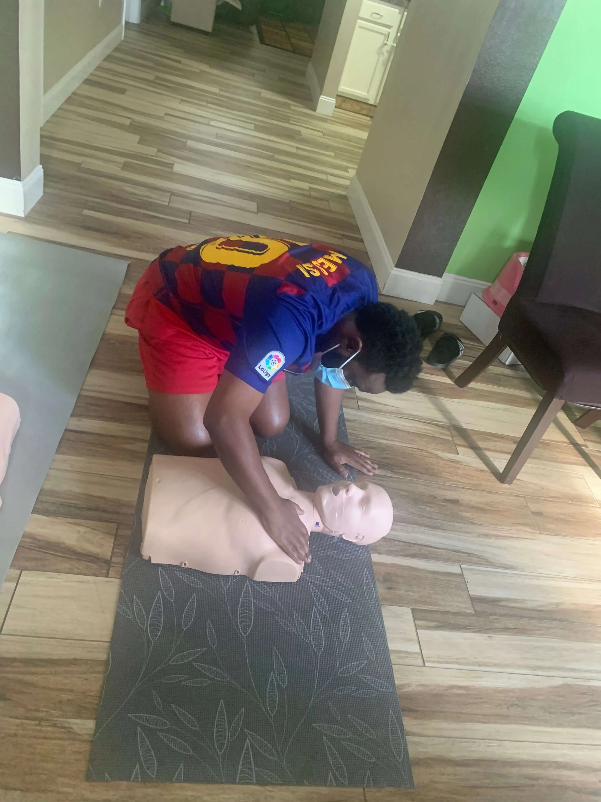 A person wearing a Barcelona soccer jersey and red shorts is practicing CPR on a mannequin lying on a mat on the wooden floor. The person is wearing a face mask and kneeling while performing chest compressions.