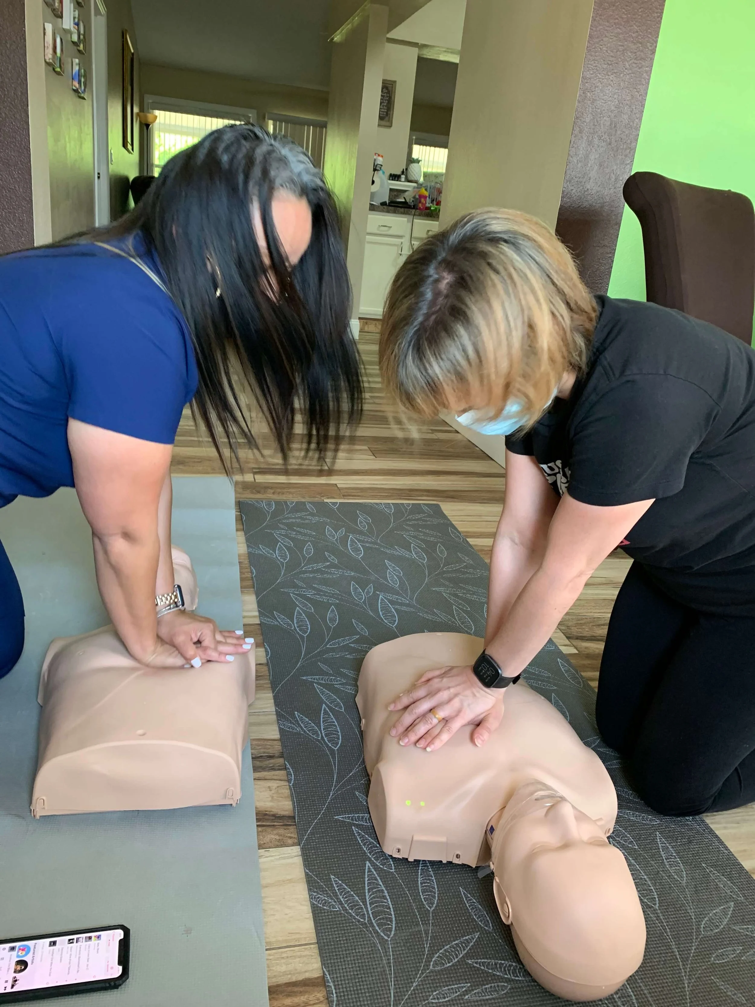 Two women practicing CPR on mannequins indoors, with a phone visible on the table nearby.