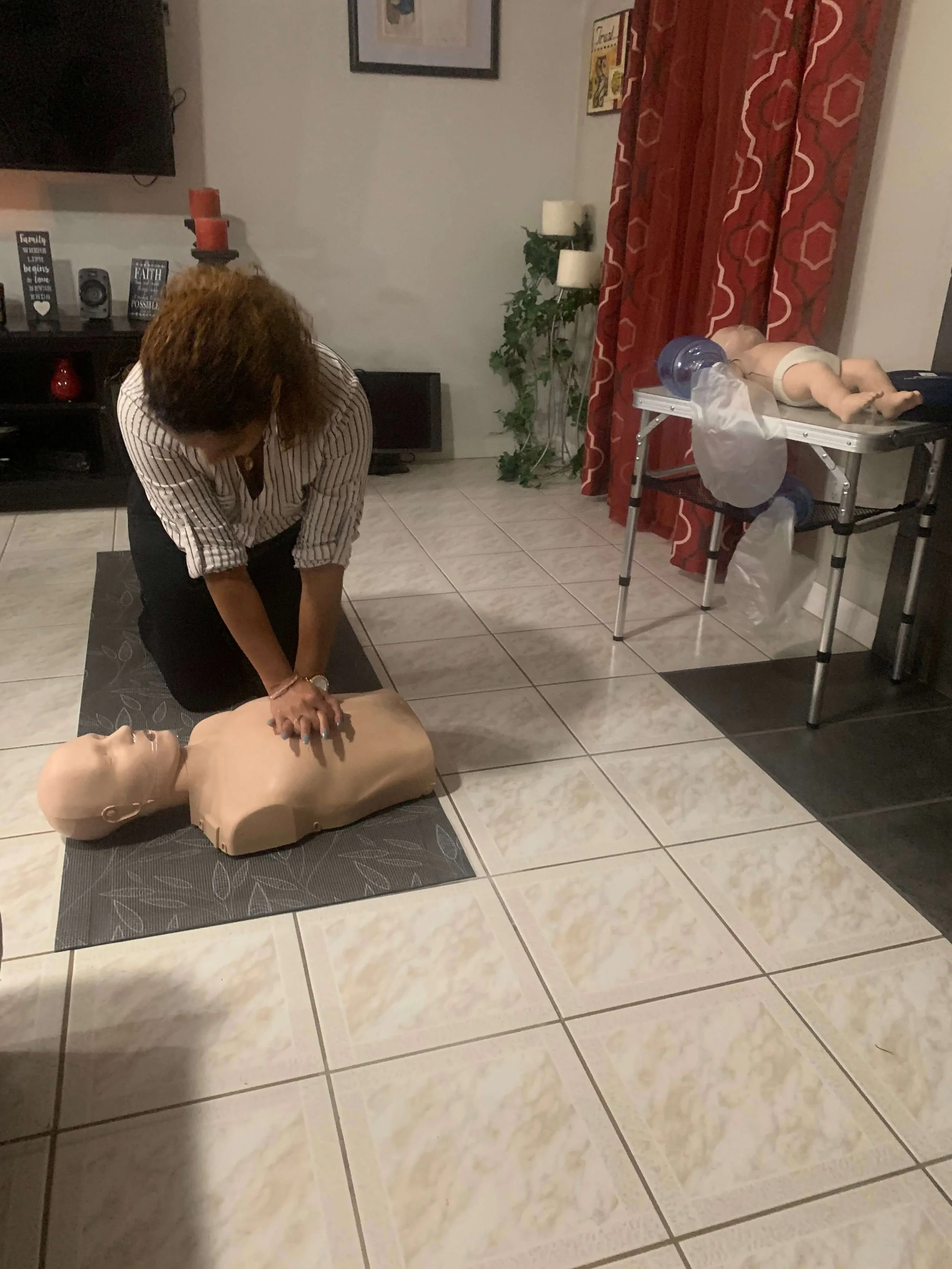 A woman practicing CPR on a medical training mannequin on the floor, with a second mannequin on a table in the background.