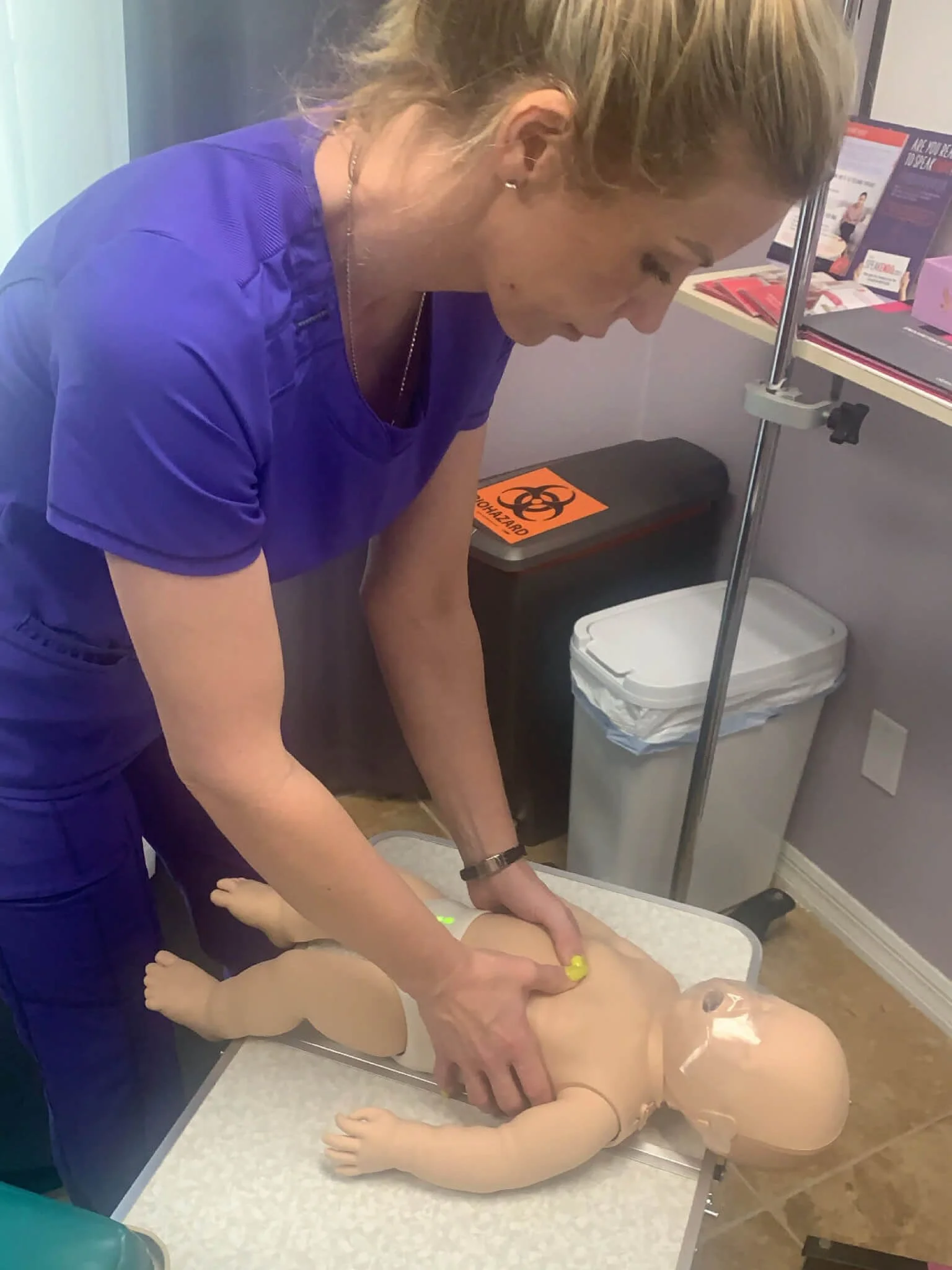 A woman in purple scrubs performing CPR on a medical training doll lying on an examination table.