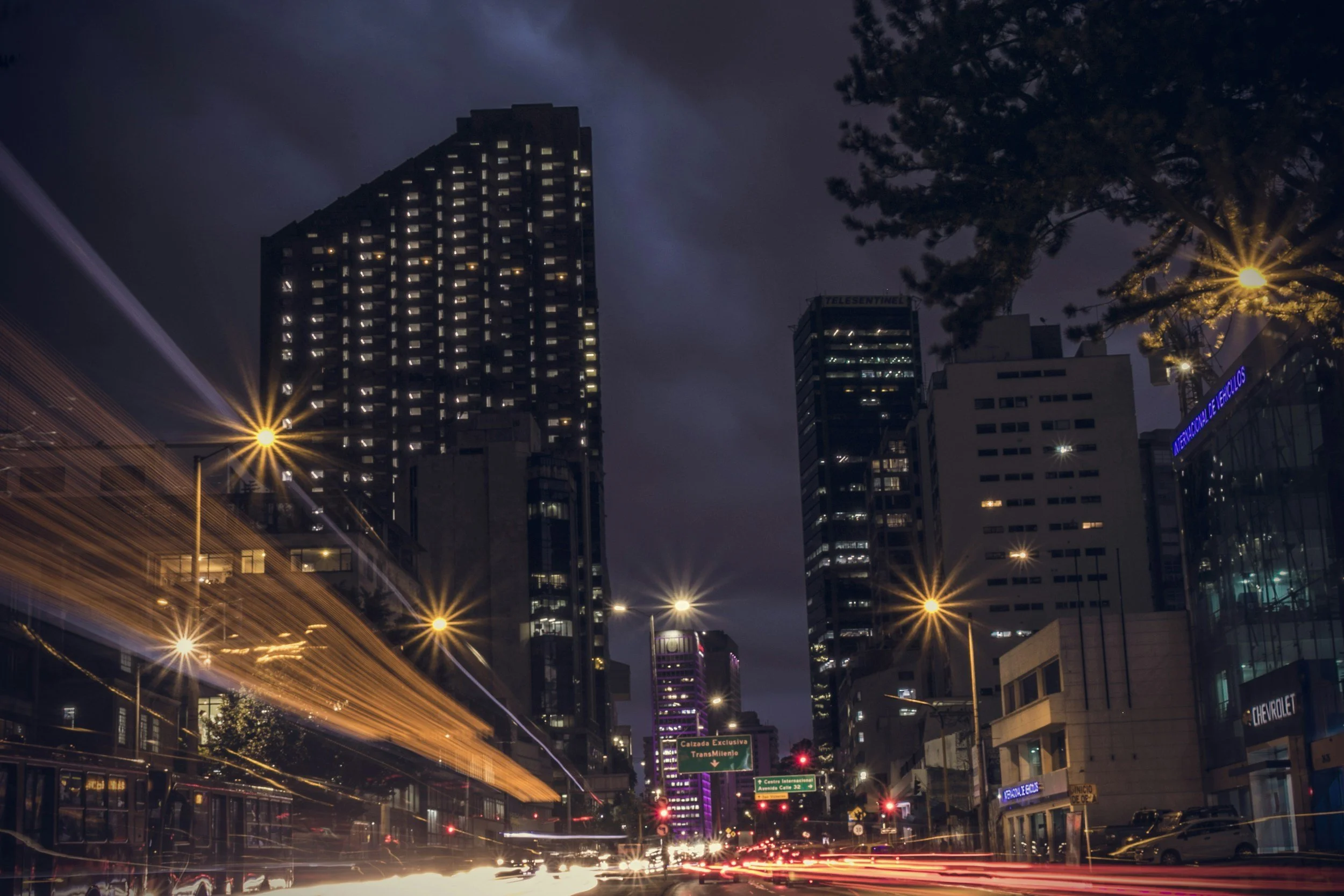 Night cityscape with tall buildings, illuminated windows, streetlights, and light trails from moving vehicles.