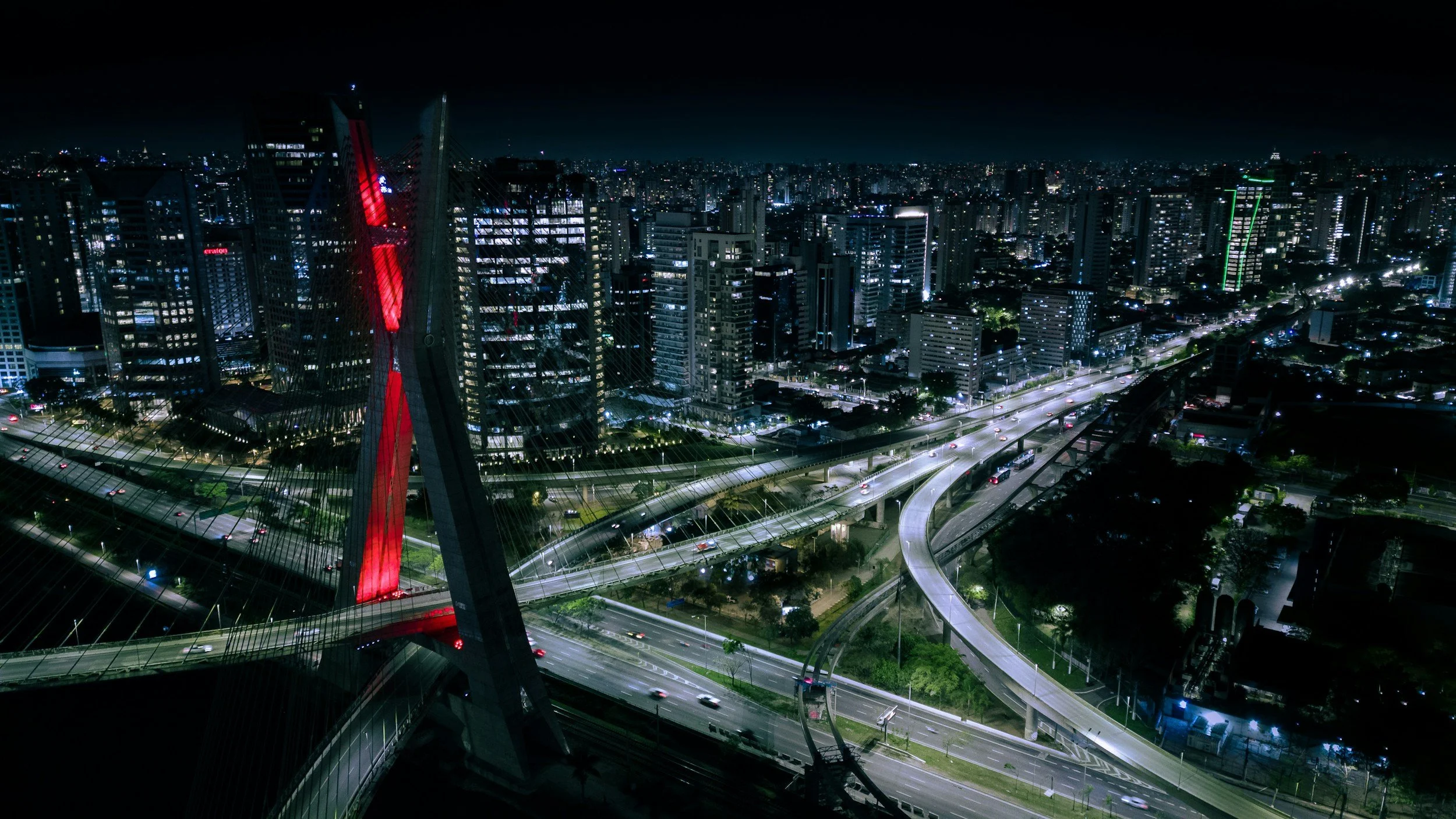 Nighttime aerial view of a cityscape with illuminated high-rise buildings, highways, and roads, with a tall structure featuring red lighting.
