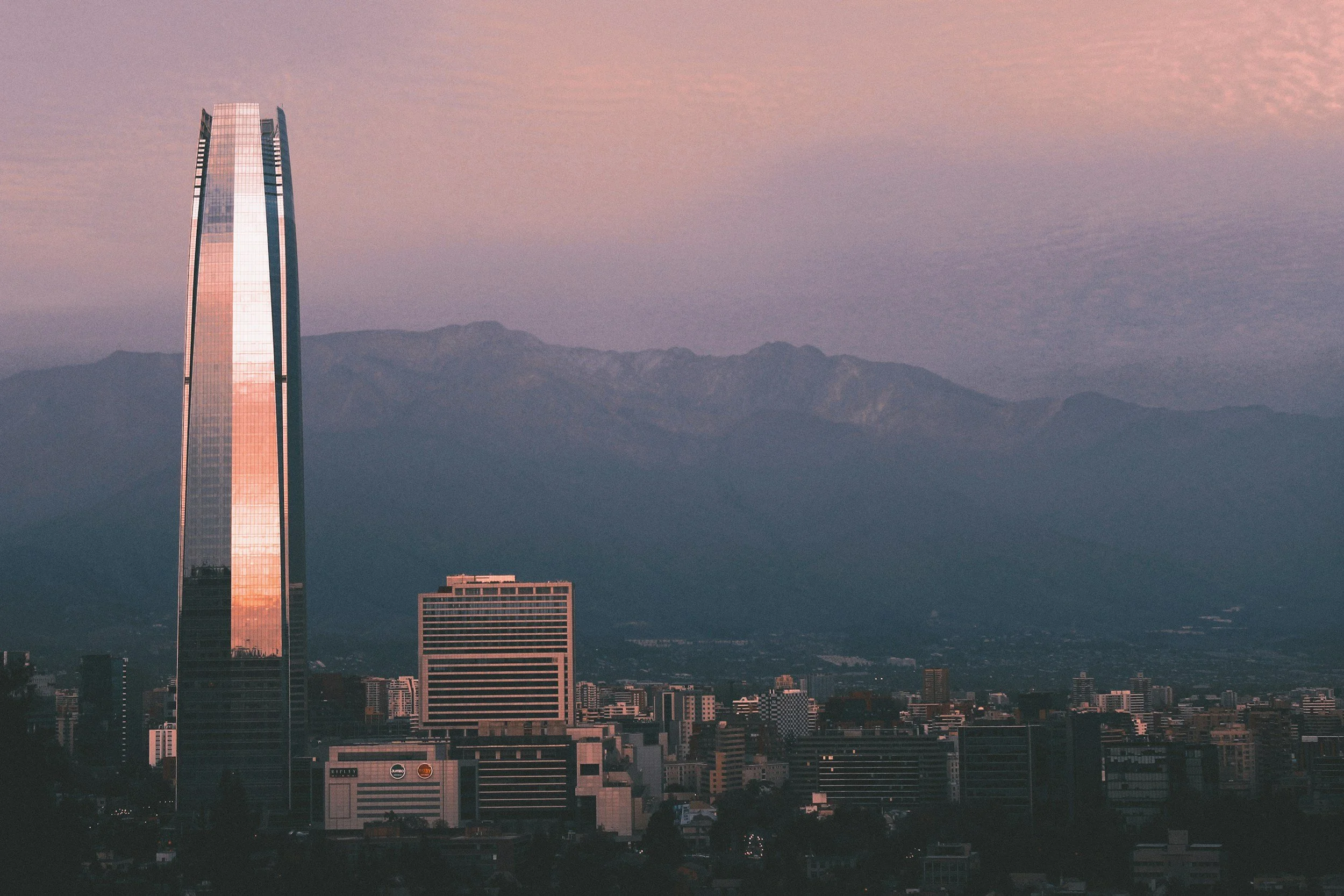 City skyline at sunset with modern skyscrapers and mountains in the background, reflecting a pinkish hue in the sky.