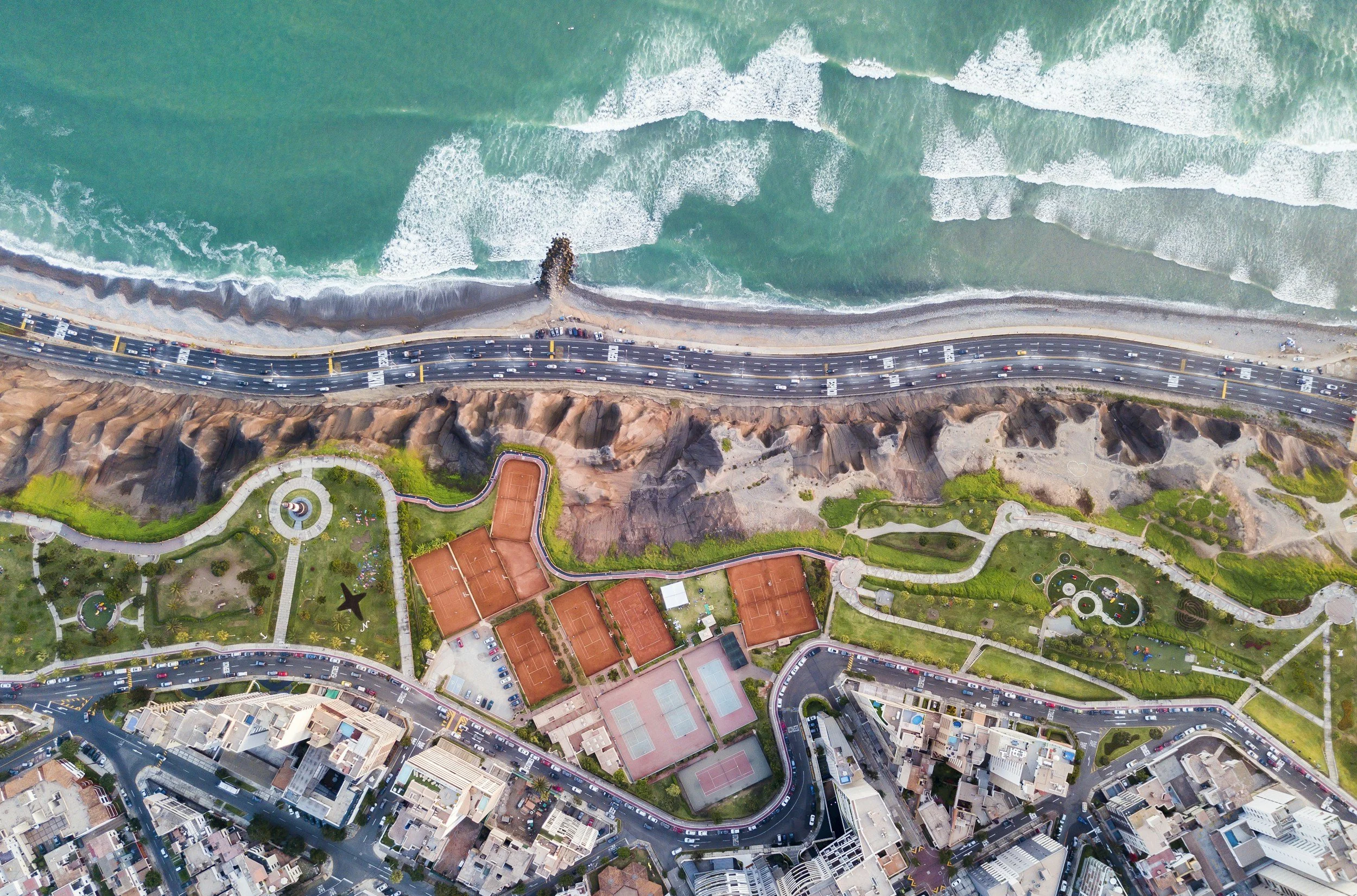 Aerial view of a coastal city showing ocean waves, a highway, tennis courts, and urban buildings.