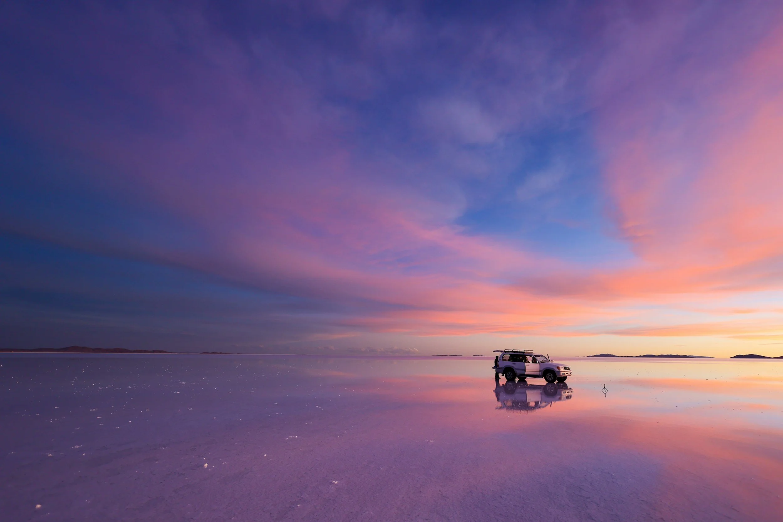 A car parked near a reflective body of water at sunset or sunrise, with colorful purple, pink, and orange sky, and distant landforms on the horizon.