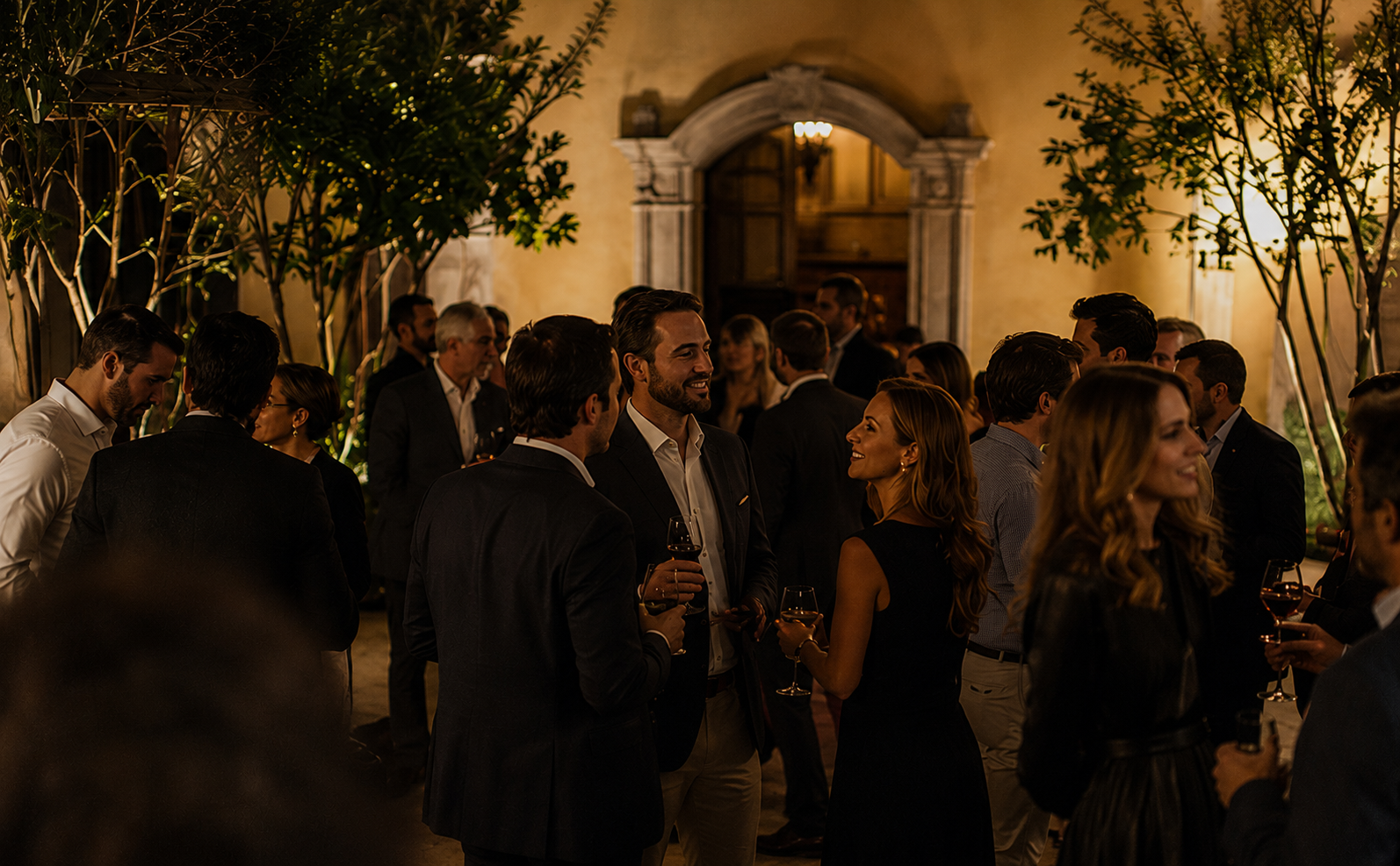 People socializing at an outdoor evening event, holding wine glasses, in a warmly lit setting with trees and an arched doorway in the background.