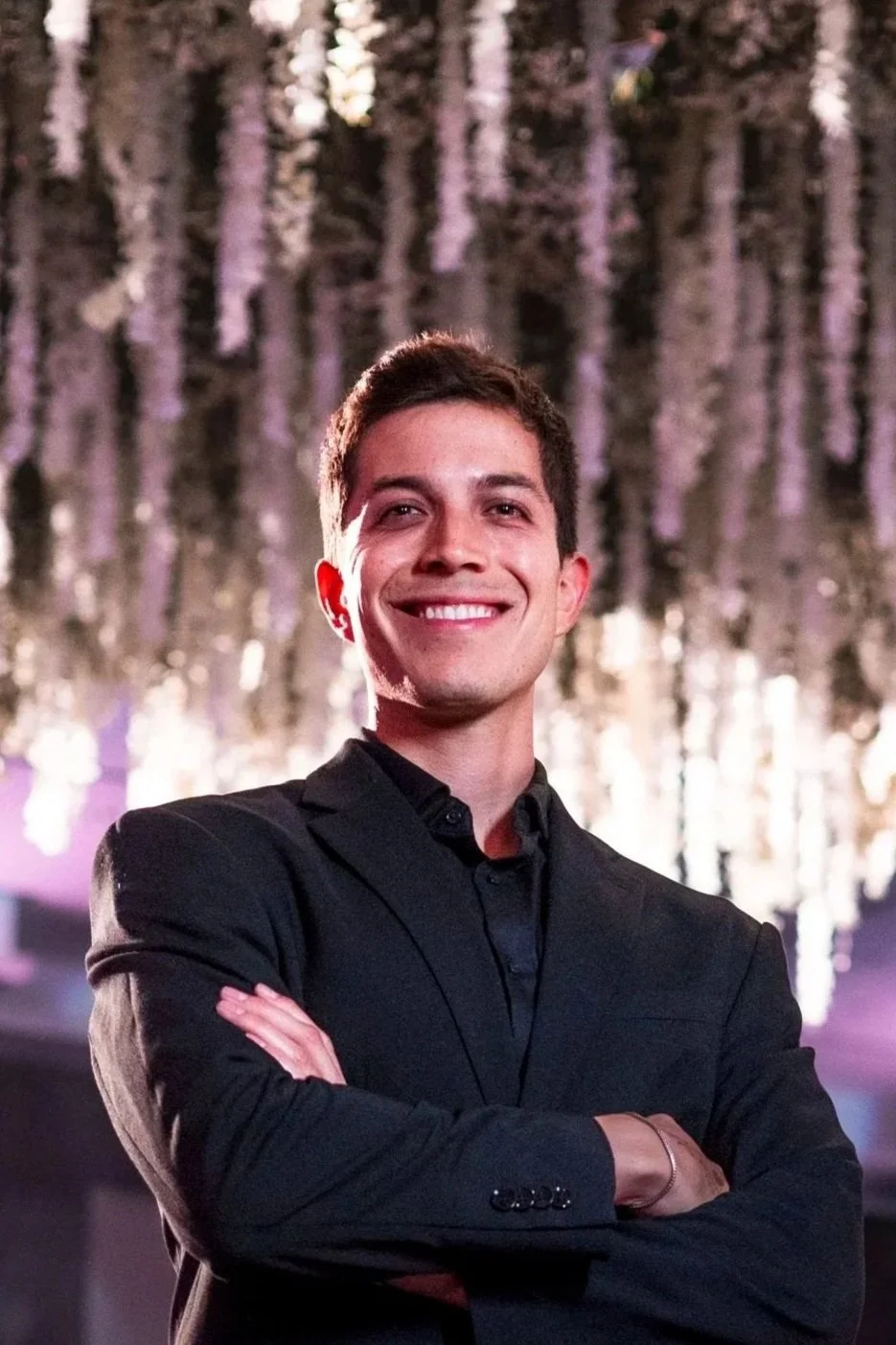 A smiling young man in a black suit with folded arms; background features a chandelier with hanging purple and white lights.