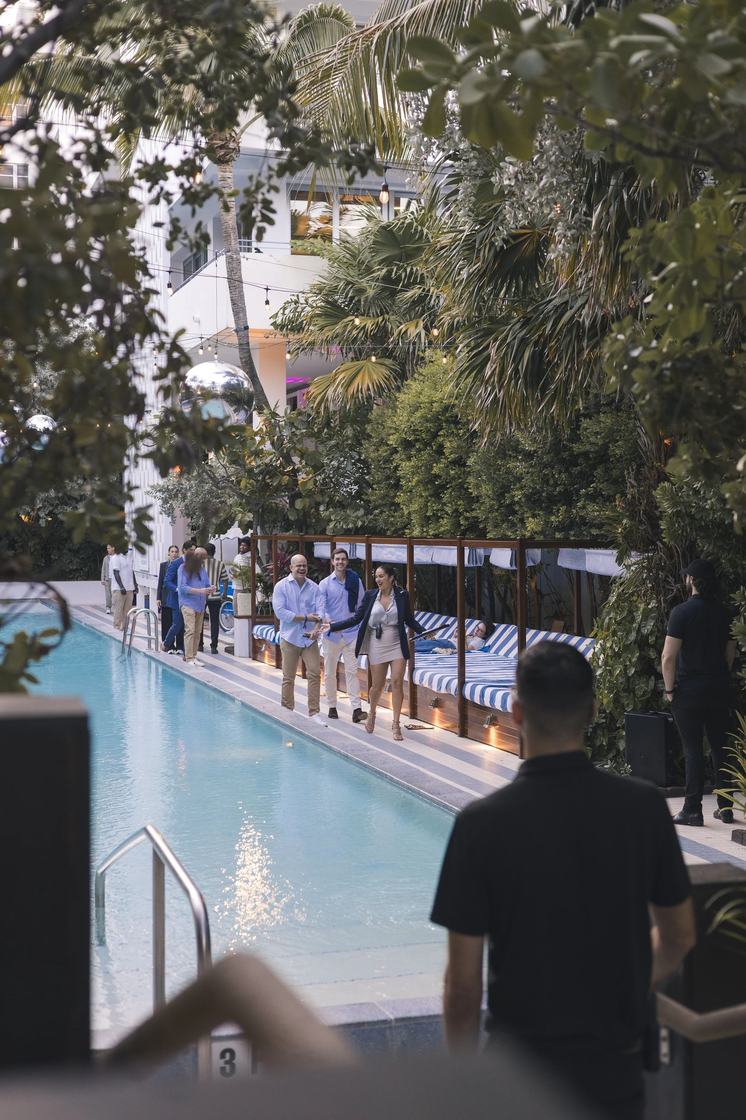 People dressed in business casual attire lined up at the edge of an outdoor swimming pool, preparing to enter the water. The area is surrounded by lush greenery, including palm trees and other tropical plants. String lights and large disco balls are hanging overhead, creating a festive atmosphere. In the background, a modern building with balconies is visible.