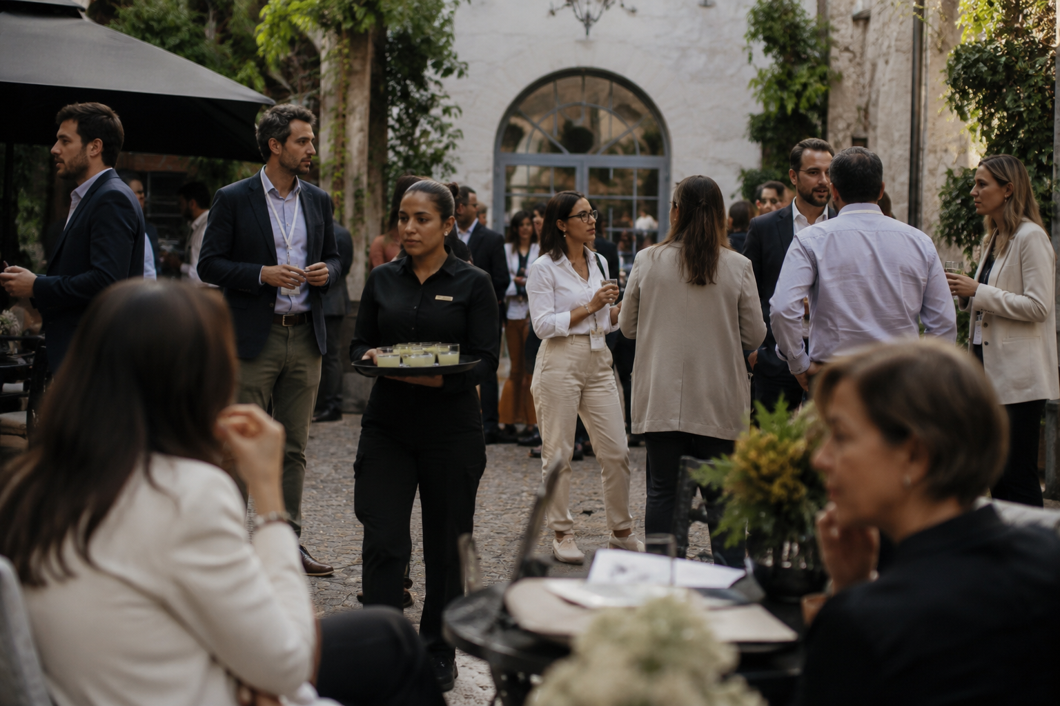 People attending a social outdoor event, some engaged in conversation, others holding drinks or trays of food, in a courtyard with greenery and a stone building.