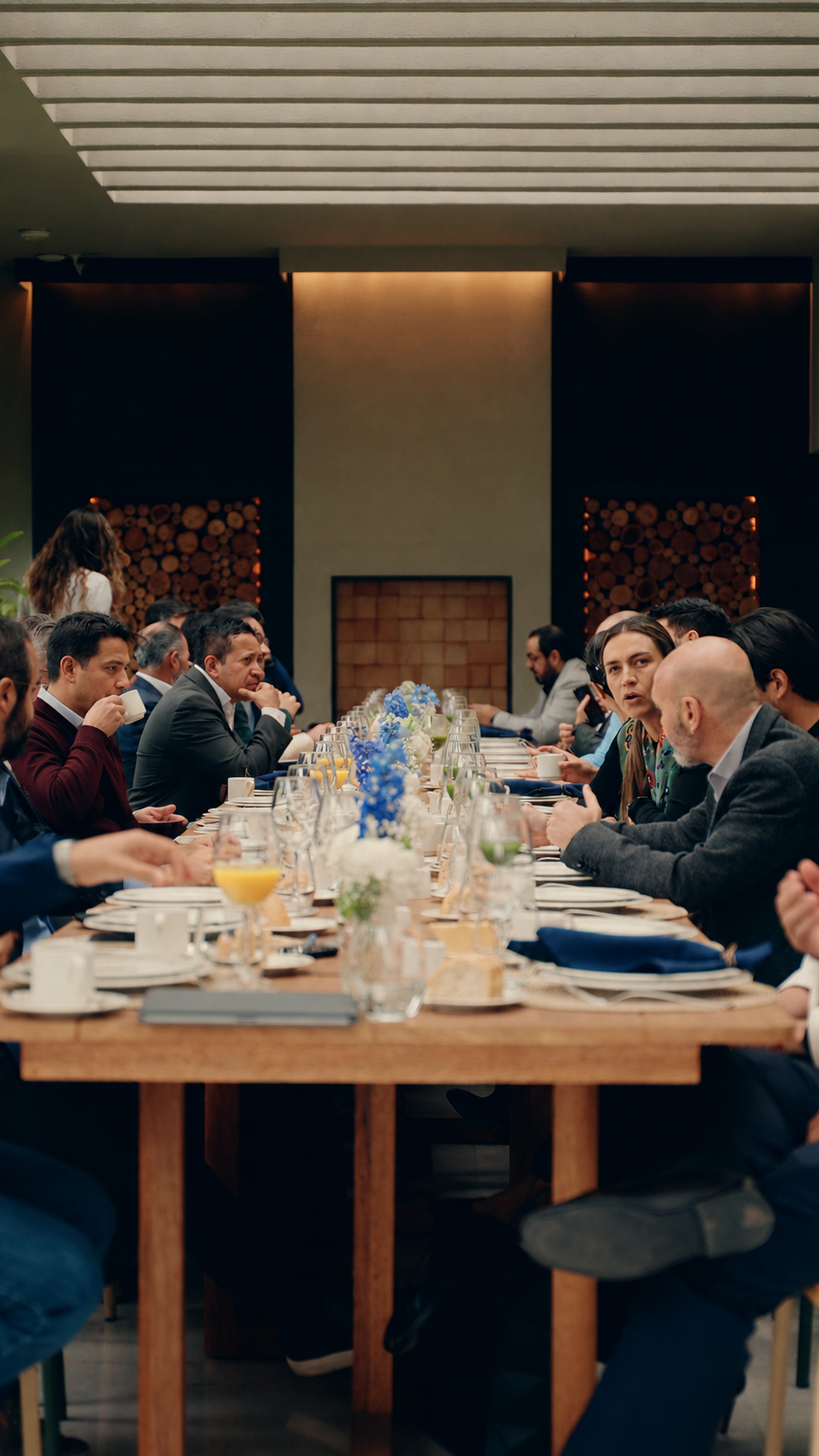 A diverse group of people seated around a long dining table in a modern restaurant, engaged in conversation, with table settings, flowers, and drinks.