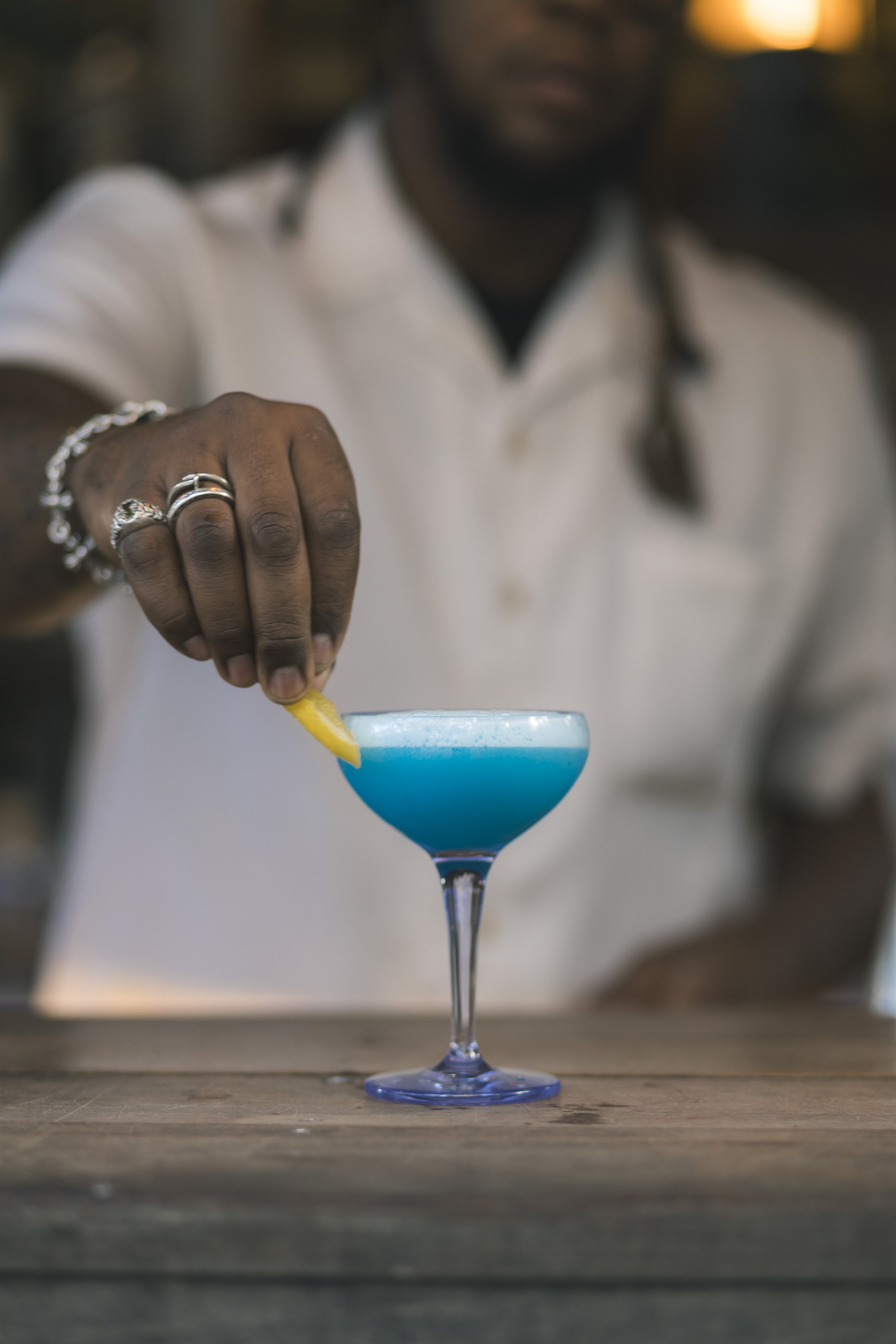 Close-up of a man's hand placing a lemon wedge into a blue cocktail in a cocktail glass on a wooden surface.