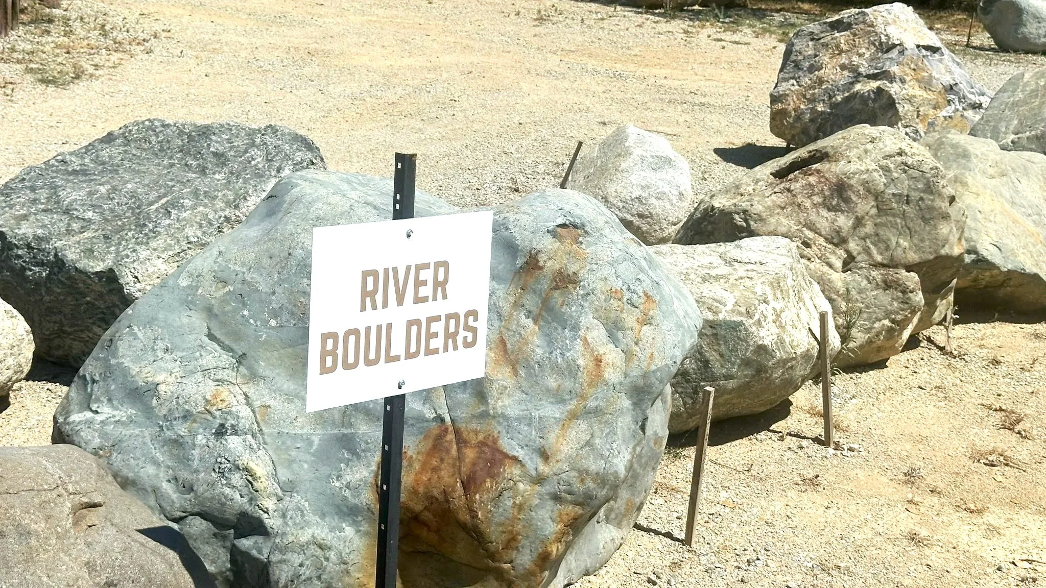 Large rocks with a sign that reads "RIVER BOULDERS," in a sandy outdoor area.