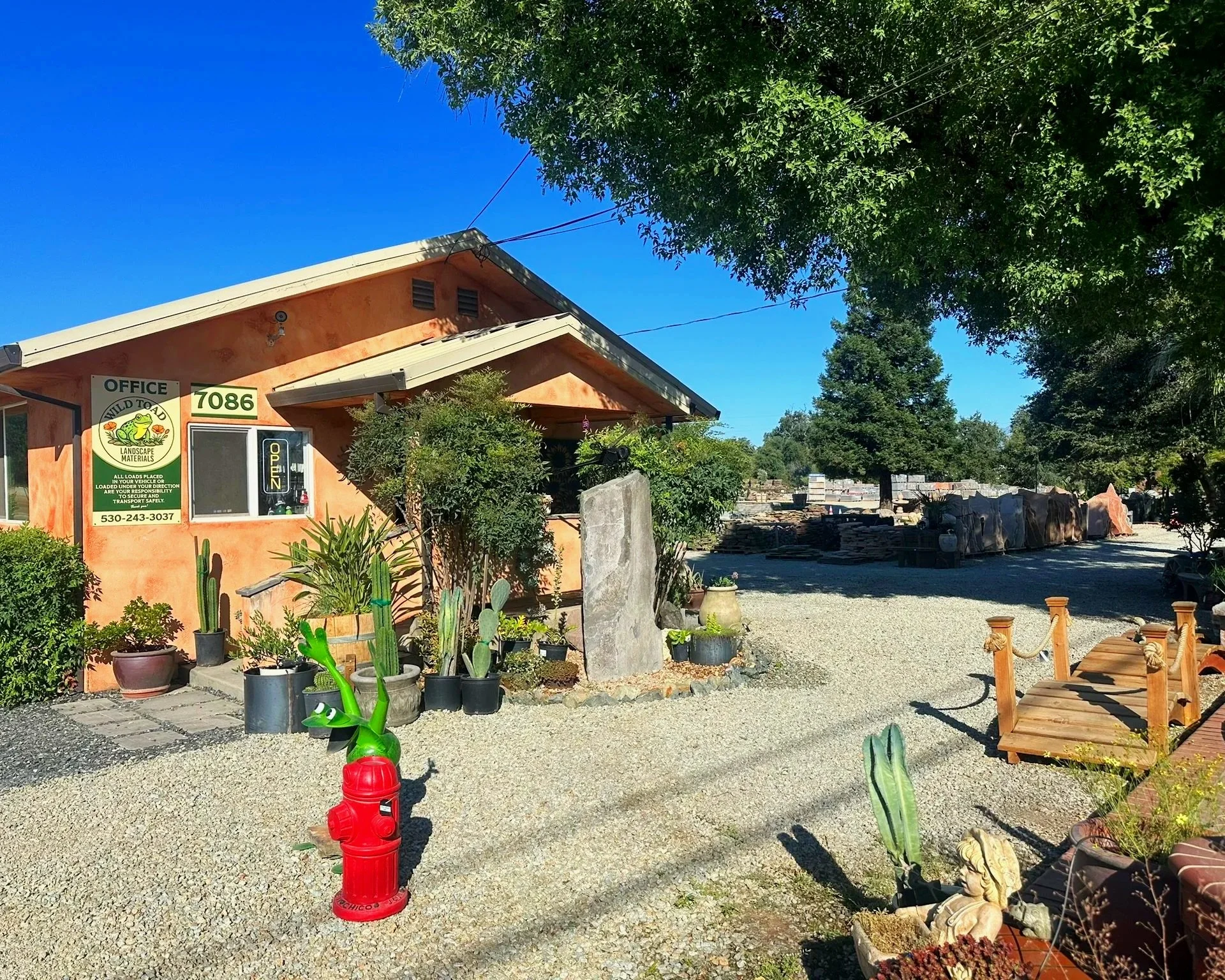 Exterior view of a small business or office at 7086 with a sign indicating landscape materials. The building has a peach-colored wall and a sign with a frog logo. There are plants and cacti in front of the building, along with gravel ground. A red fire hydrant is visible in the foreground. The background features a clear blue sky and trees.