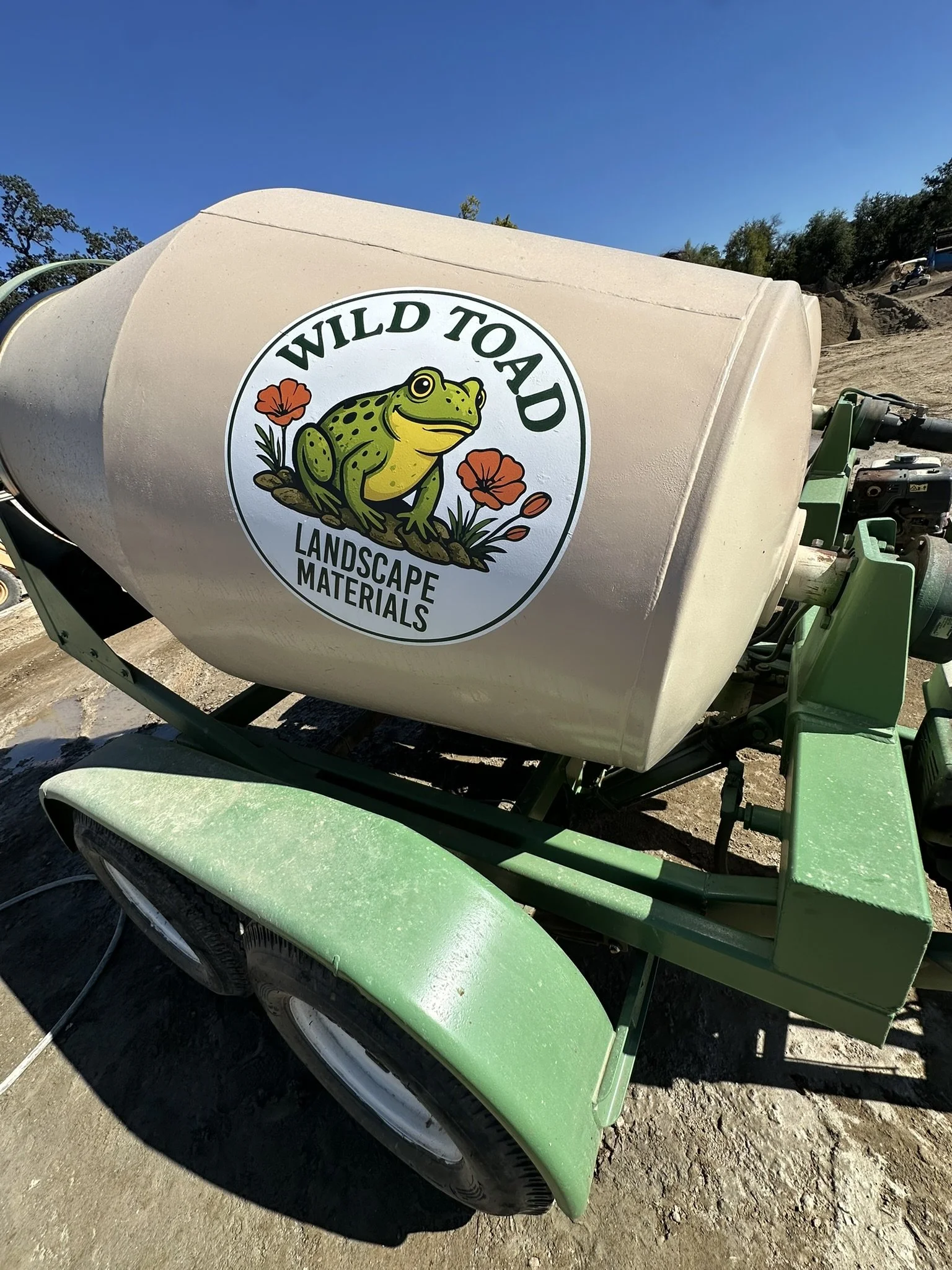 A cement mixer truck with a logo of a frog sitting on a rock among flowers, with the words "Wild to Aid" and "Landscape Materials" on the side, on a sunny outdoor construction site.