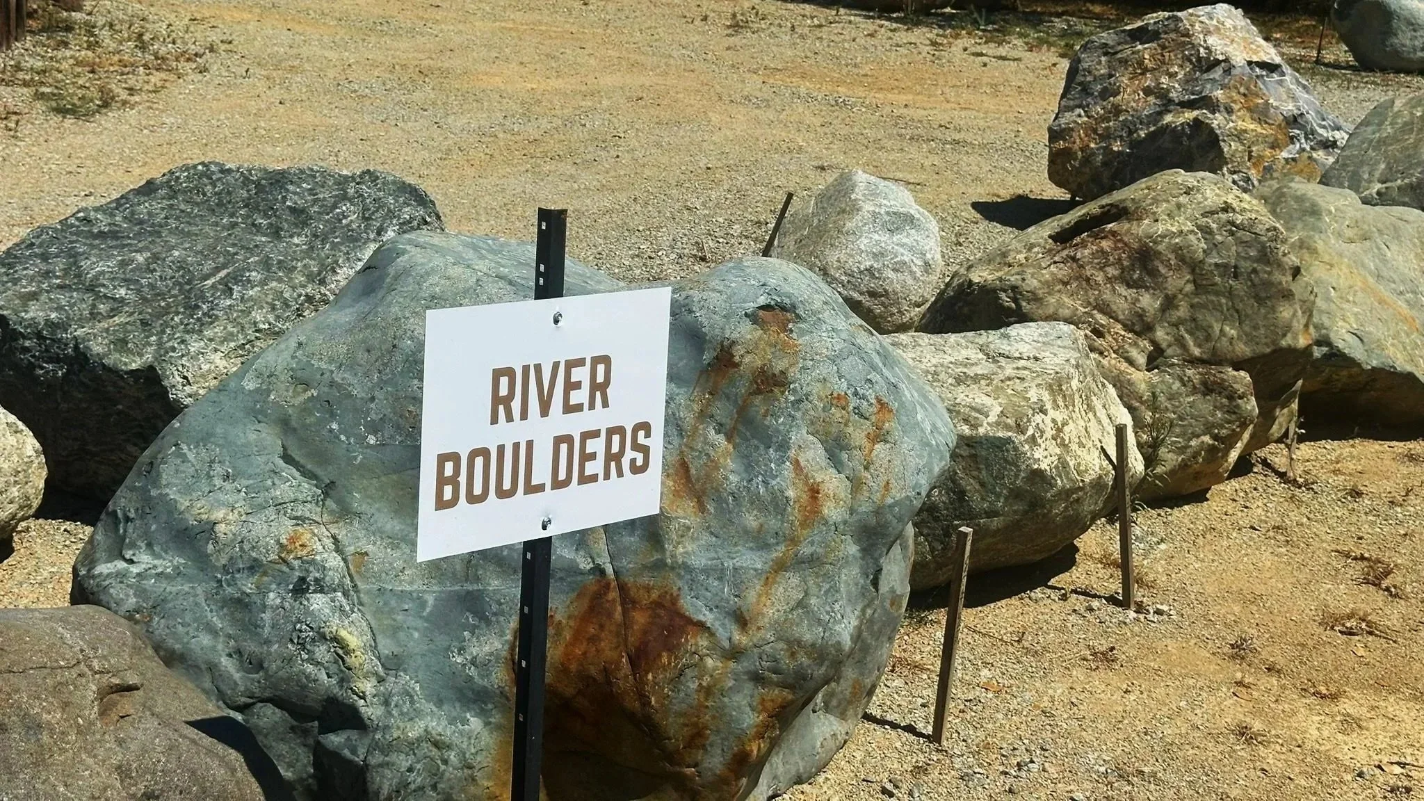 A collection of large rocks with a sign that reads 'River Boulders' in front of them, situated in a sandy outdoor area.