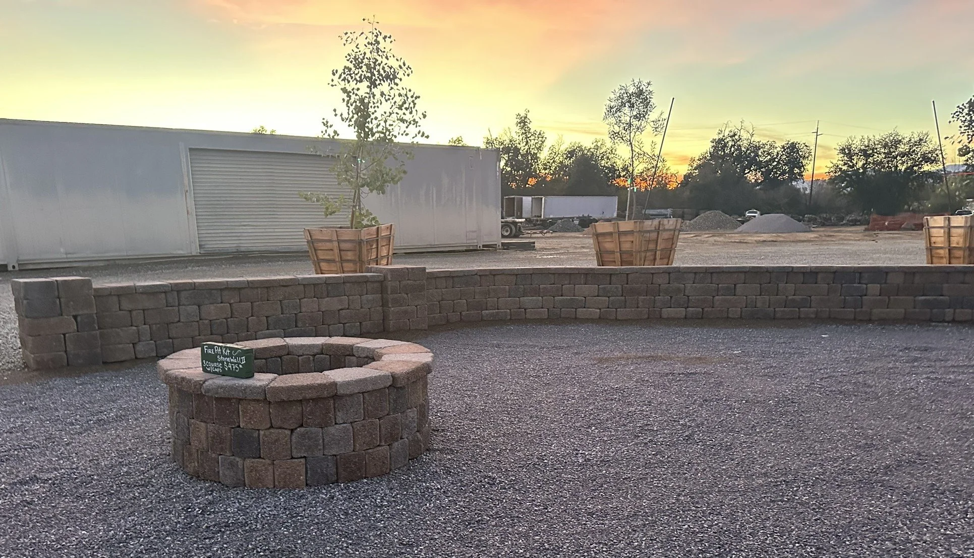 Outdoor scene at sunset featuring a circular brick fire pit with a small chalkboard sign, a curved brick wall, two large wooden planters with young trees, and gravel ground, with trucks and trees in the background.