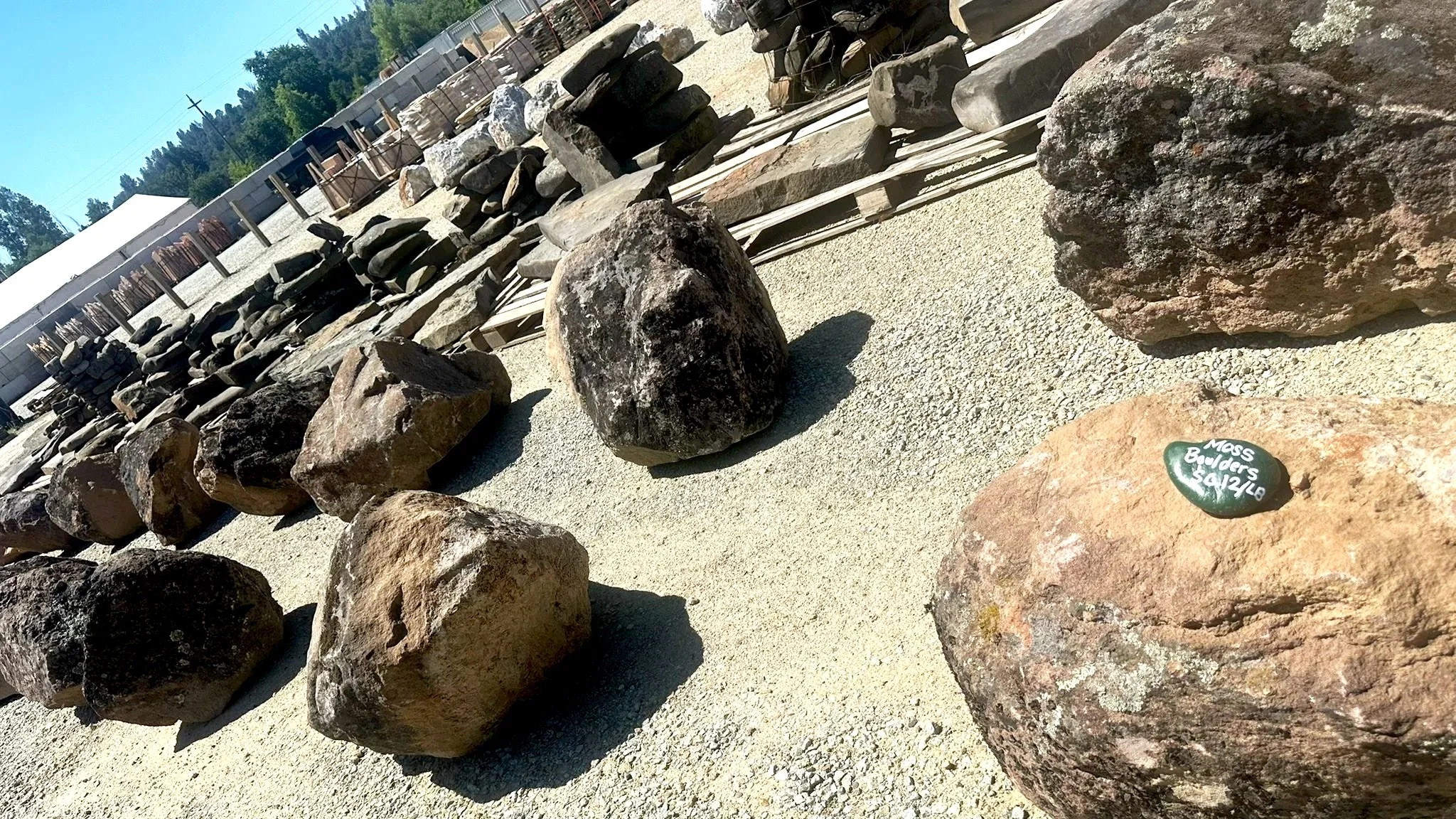 Various large moss boulders rocks displayed outdoors with a small green stone inscribed 'Mass Boulders 9/12/16'. In the background, there are stacks of smaller rocks and a white canopy or tent, with trees and a utility pole visible in the distance.