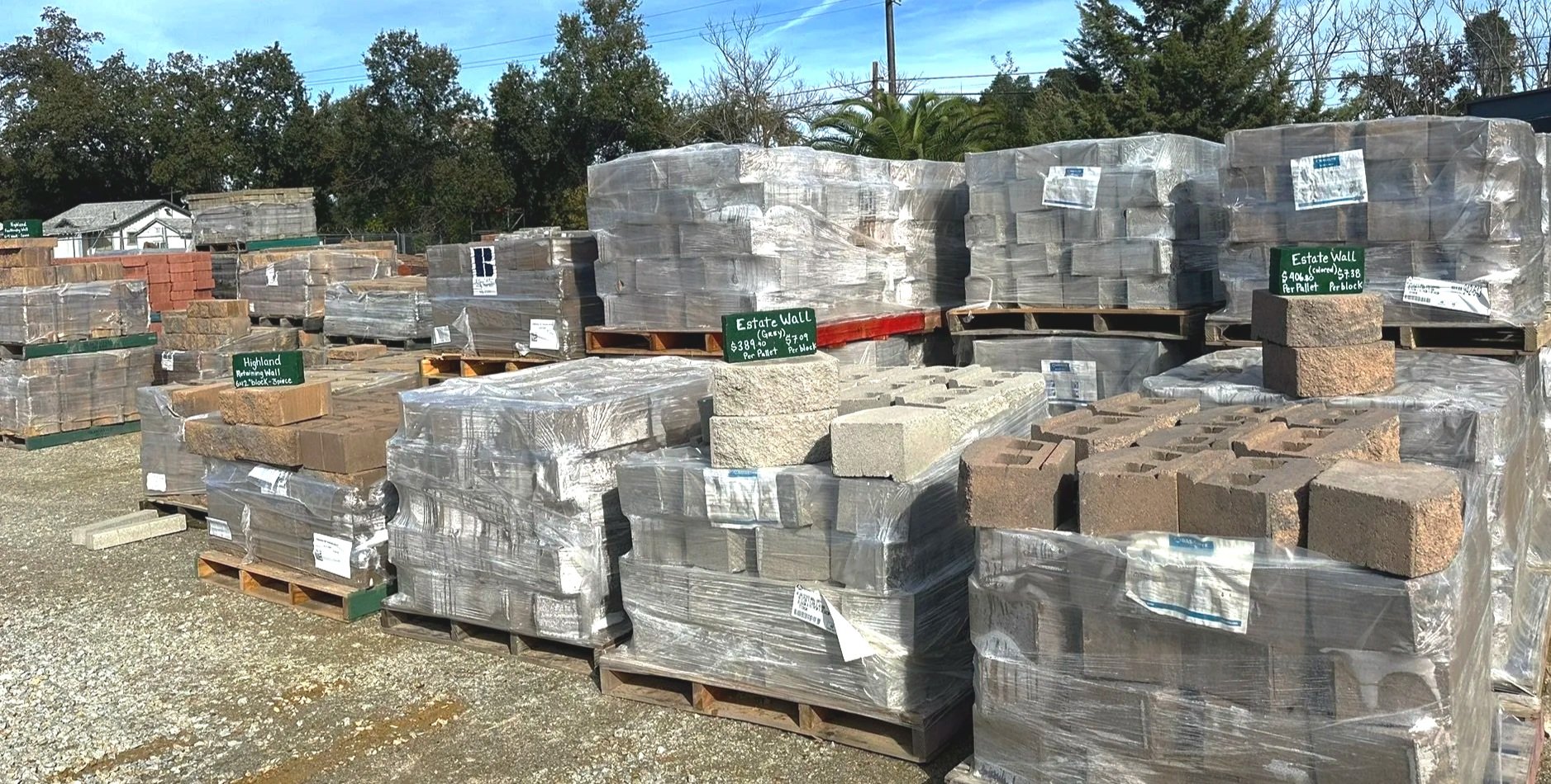 Stacks of building bricks arranged on pallets outdoors, with green signs indicating prices and types, some wrapped in plastic, with trees and a house in the background.