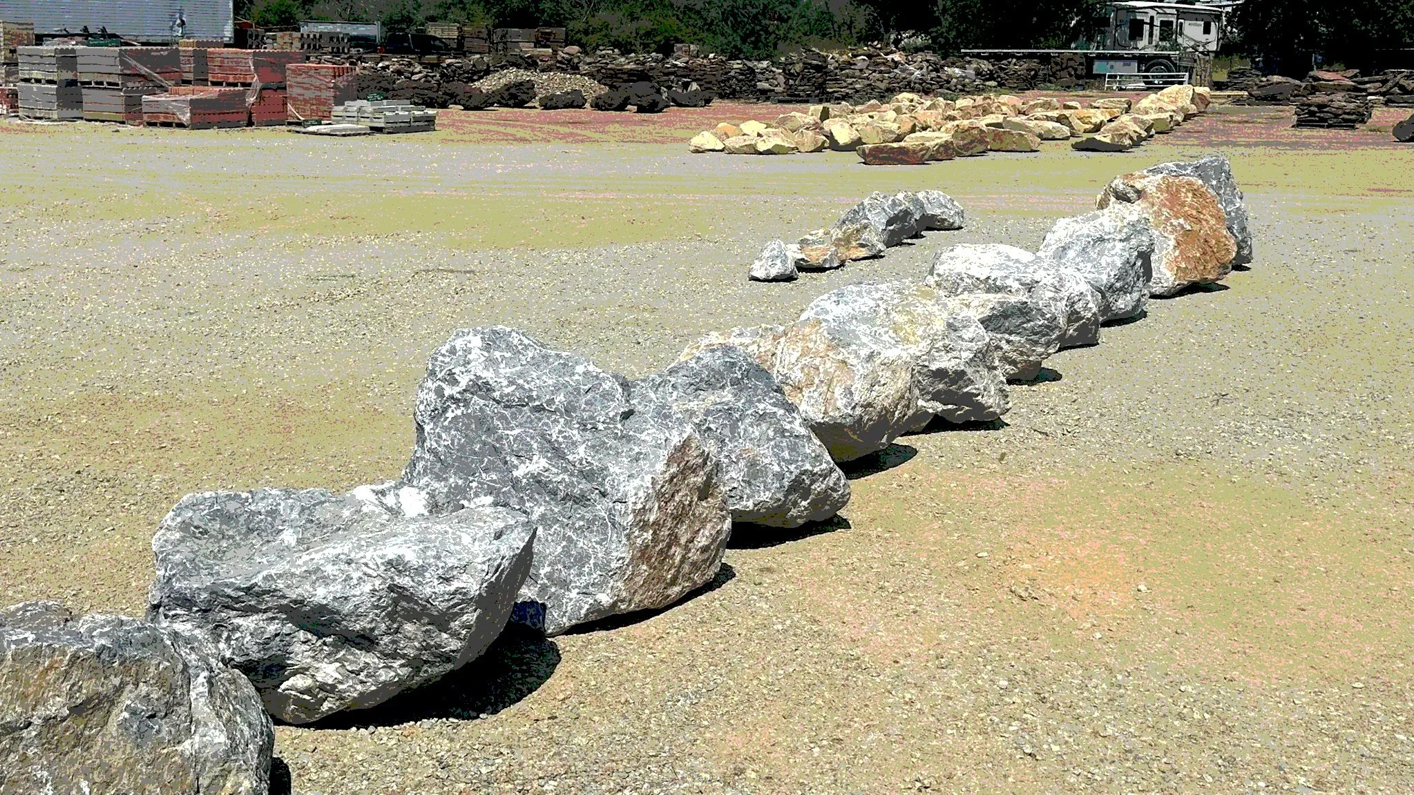 A row of large limestone boulder rocks on a sandy surface at a construction site with buildings and trees in the background.