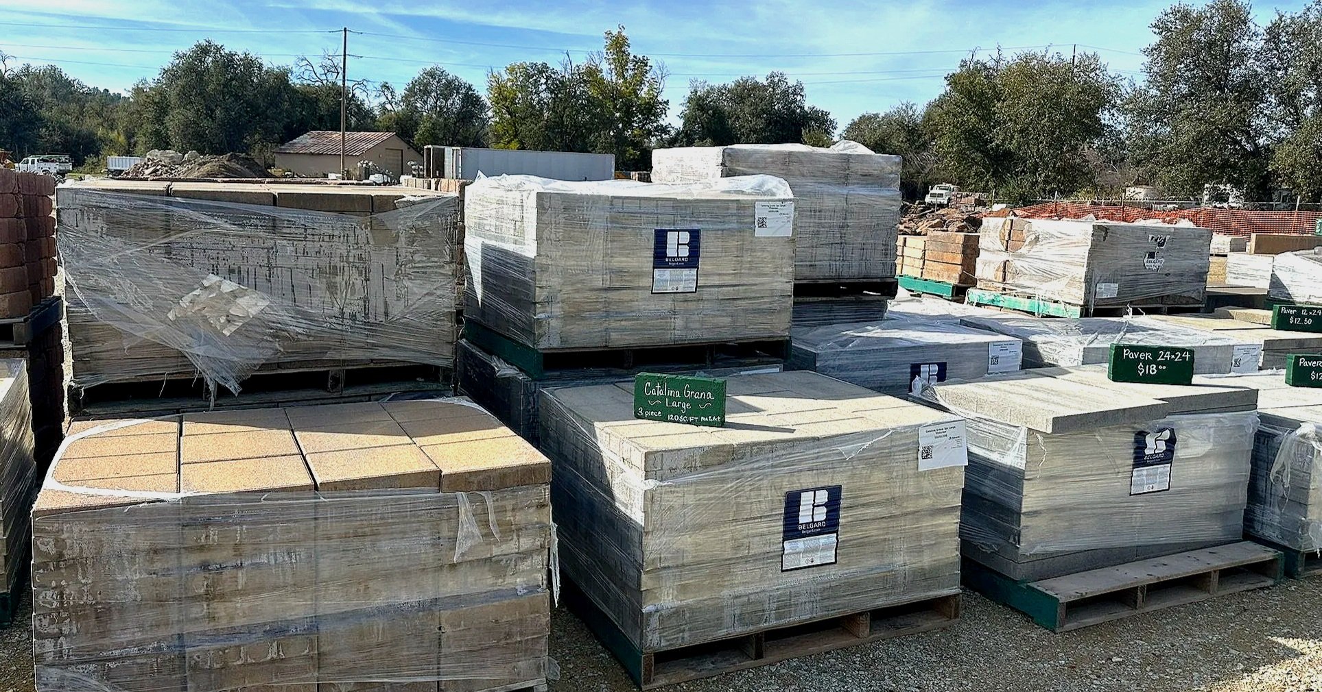 Outdoor display of large stone and tile slabs wrapped in plastic at a building materials store. Various labels and prices are visible on the slabs, with trees and utility poles in the background.