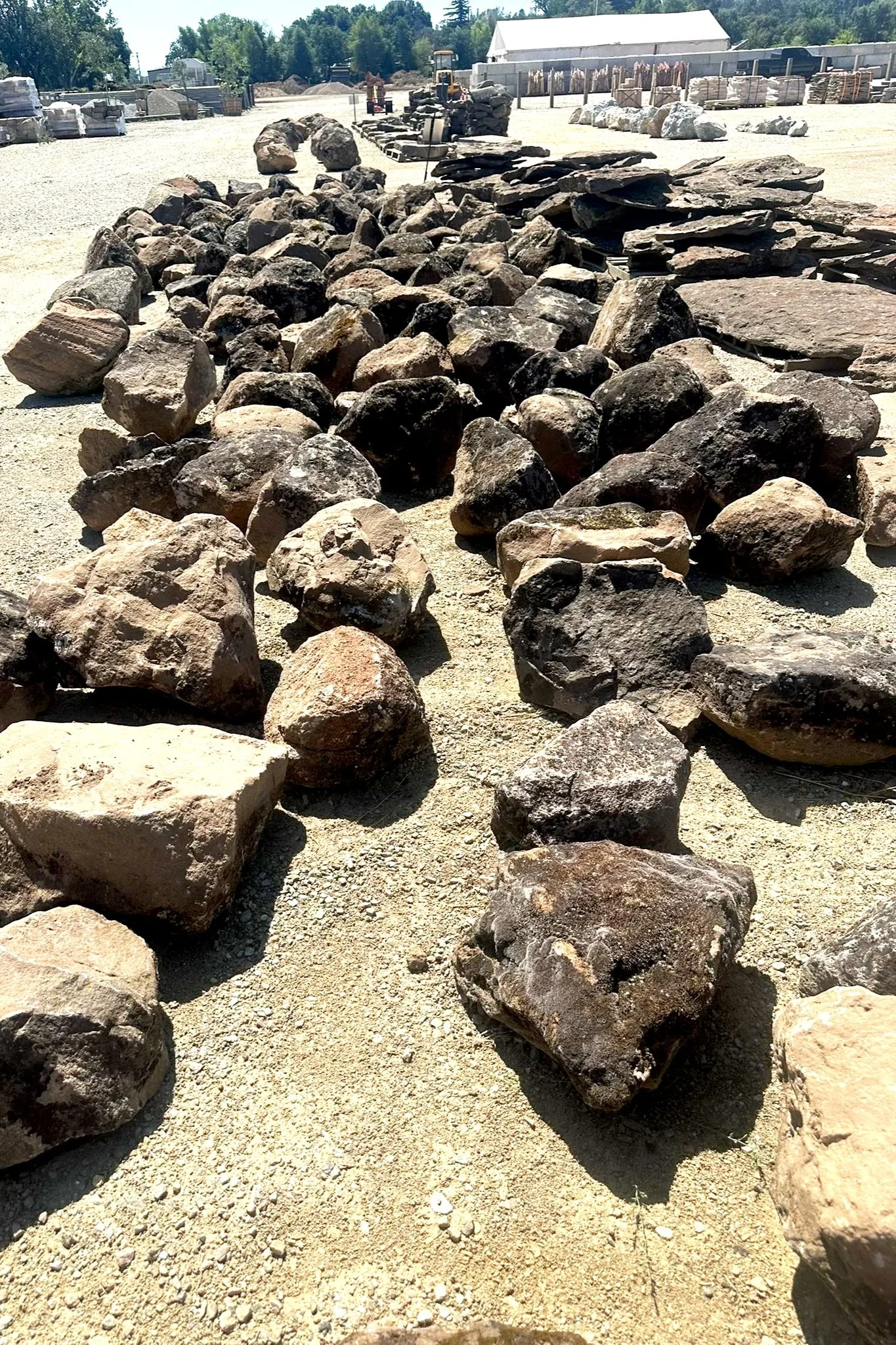 A gravel yard with large Moss rocks and stones arranged in rows, with some pallets of stone materials and a small construction vehicle in the background.