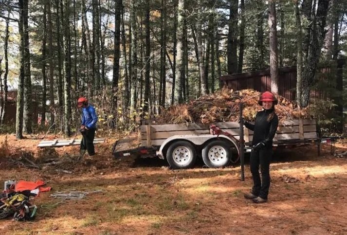 Two workers wearing safety helmets stand near a trailer filled with tree branches and debris in a wooded area.