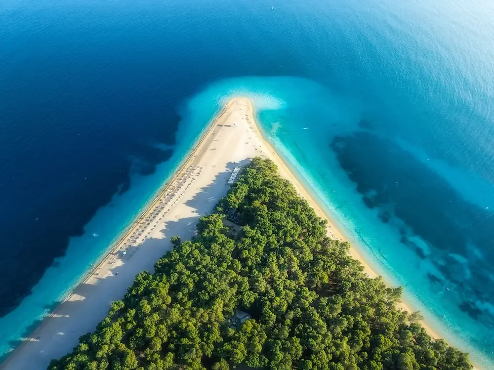 Aerial view of a narrow, elongated island with a large green forest area, sandy beaches on both sides, and a parking area at the far end, surrounded by deep blue ocean water.
