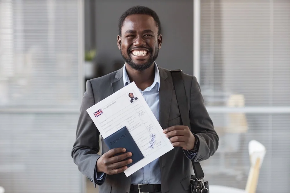 A smiling man in a suit holding a passport, a travel document, and a 'approved' stamp in an office setting.