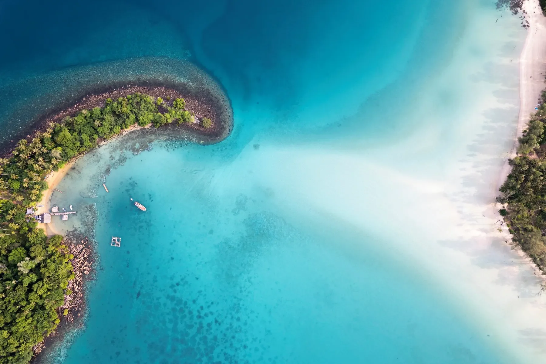 Aerial view of a boat near a small island with lush green trees, surrounded by turquoise ocean waters with varying shades, rocky shoreline, and sandy beaches.