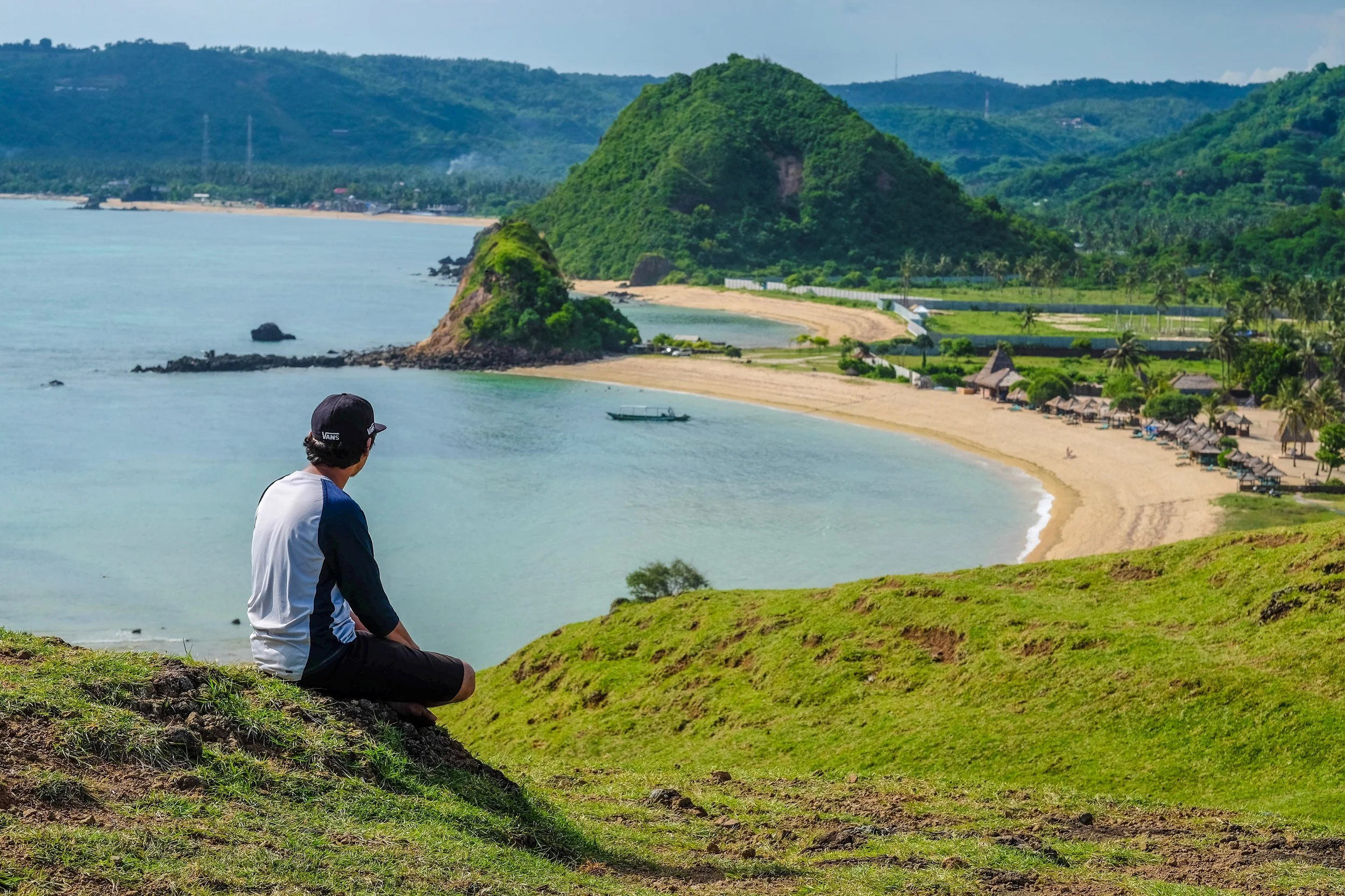 A person sitting on a grassy hill overlooking a tropical beach, with mountains and greenery in the background.