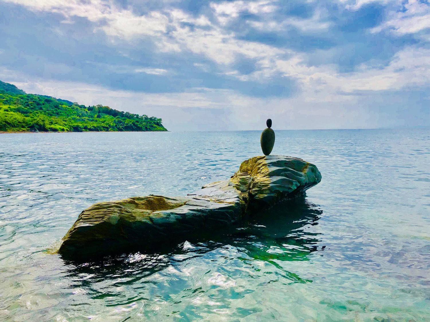 A rock formation in the water with a stacked stone sculpture on top, near a grassy, tree-covered shoreline under a partly cloudy sky.