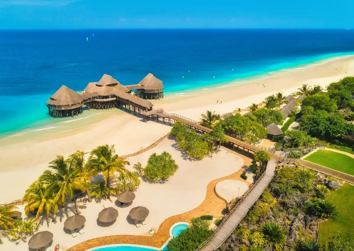 Aerial view of a tropical beach resort with overwater bungalows connected by a wooden walkway, surrounded by palm trees, sandy beaches, and clear blue ocean waters.