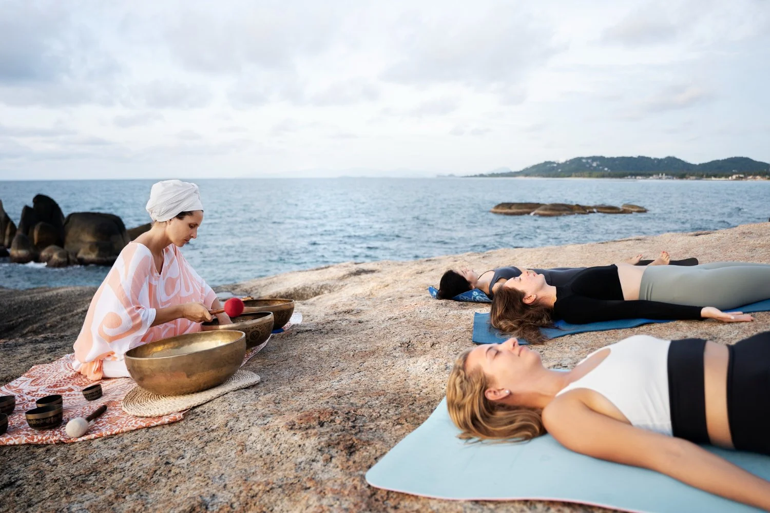 Women lying on yoga mats on a rocky beach, practicing yoga or meditation, as an instructor plays singing bowls nearby with the ocean and mountains in the background.