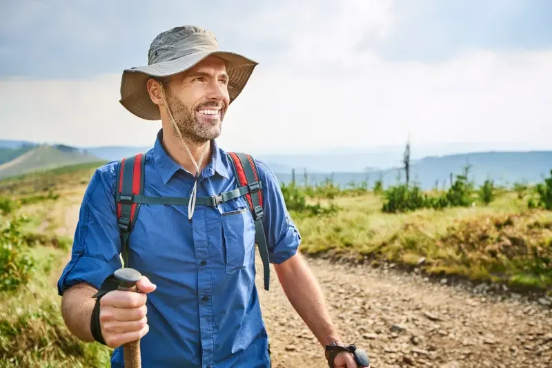 Smiling man wearing a hat and blue shirt, hiking on a trail with a backpack and trekking poles, in a scenic outdoor landscape with hills and a cloudy sky.
