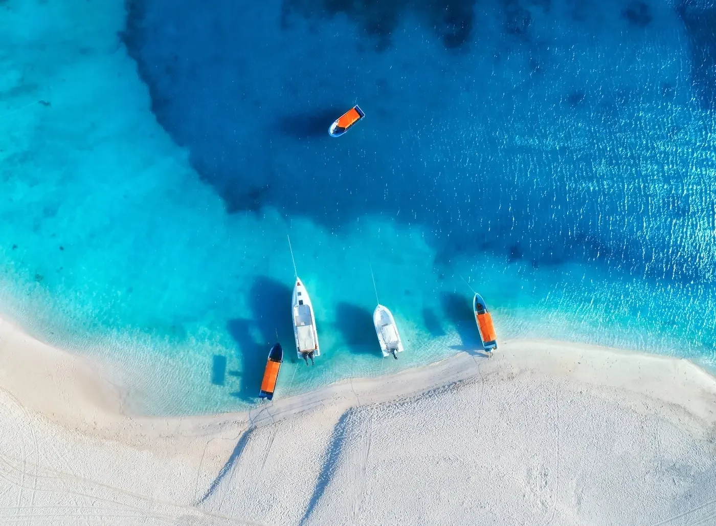 Aerial view of three sailboats anchored near a sandy beach, with clear blue water and a boat drifting further out to sea.
