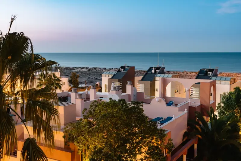 View of hotel rooftops with the ocean in the background during sunset or twilight, including palm trees and a clear sky.