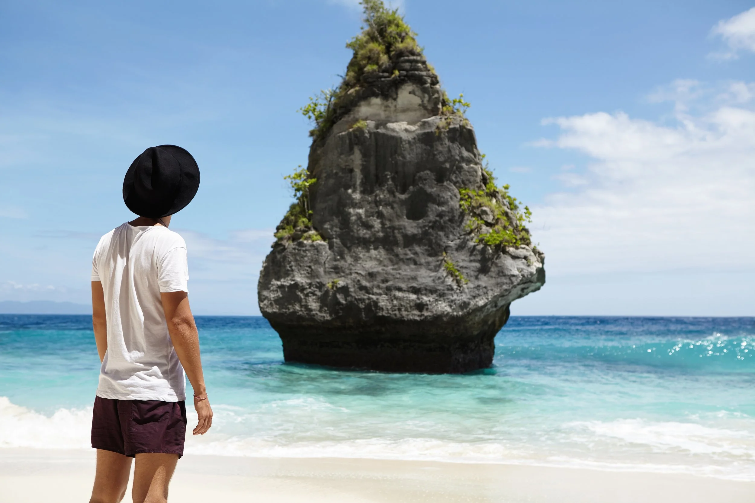 Person wearing a white t-shirt, maroon shorts, and a black hat stands on a sandy beach facing the ocean, with a large rock formation in the water nearby under a partly cloudy sky.