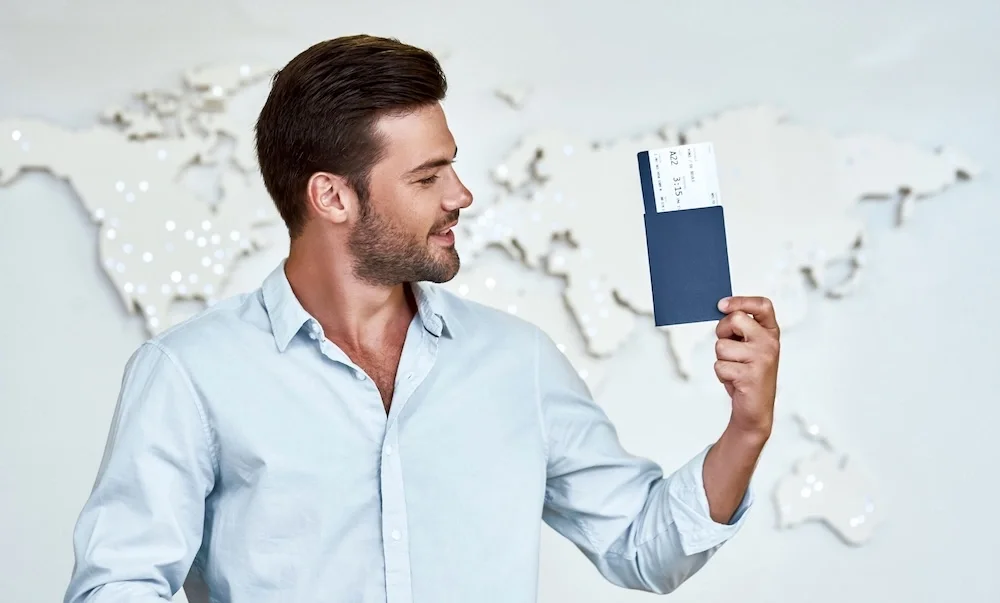 A man holding a passport with boarding passes inside in front of a world map wall display.