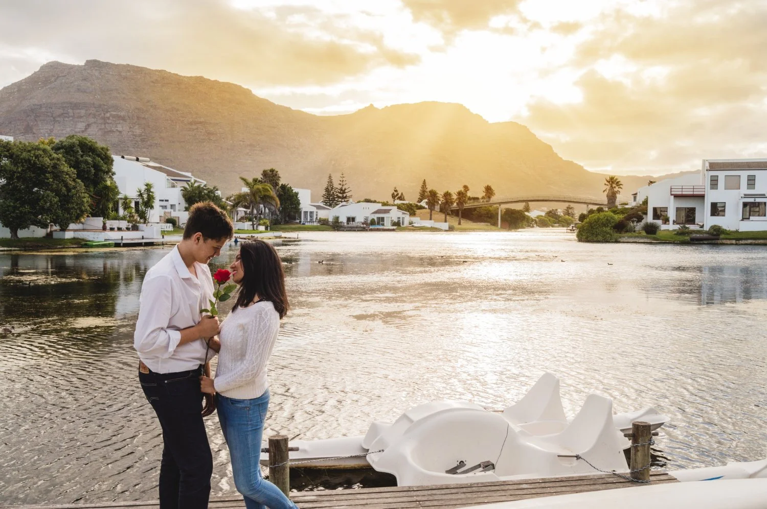 A couple on a dock by a river at sunset, with the man holding a single red rose and the woman smiling at him, overlooking white buildings and mountains in the background.