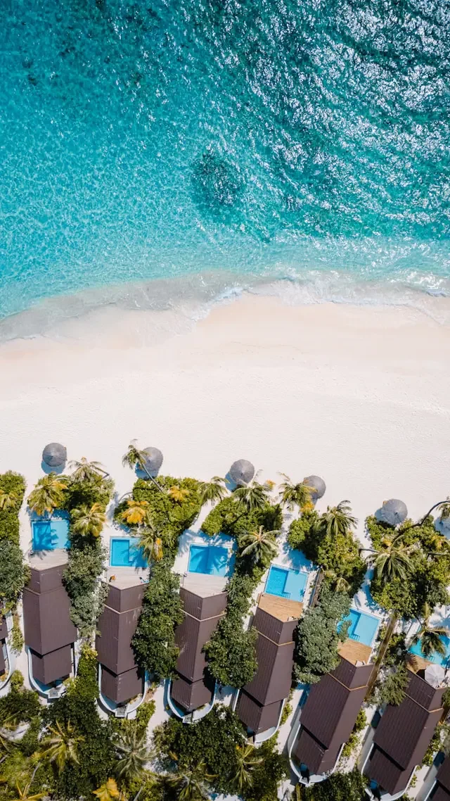 Aerial view of beachside resort with swimming pools, palm trees, and overwater bungalows near a sandy beach and turquoise ocean.