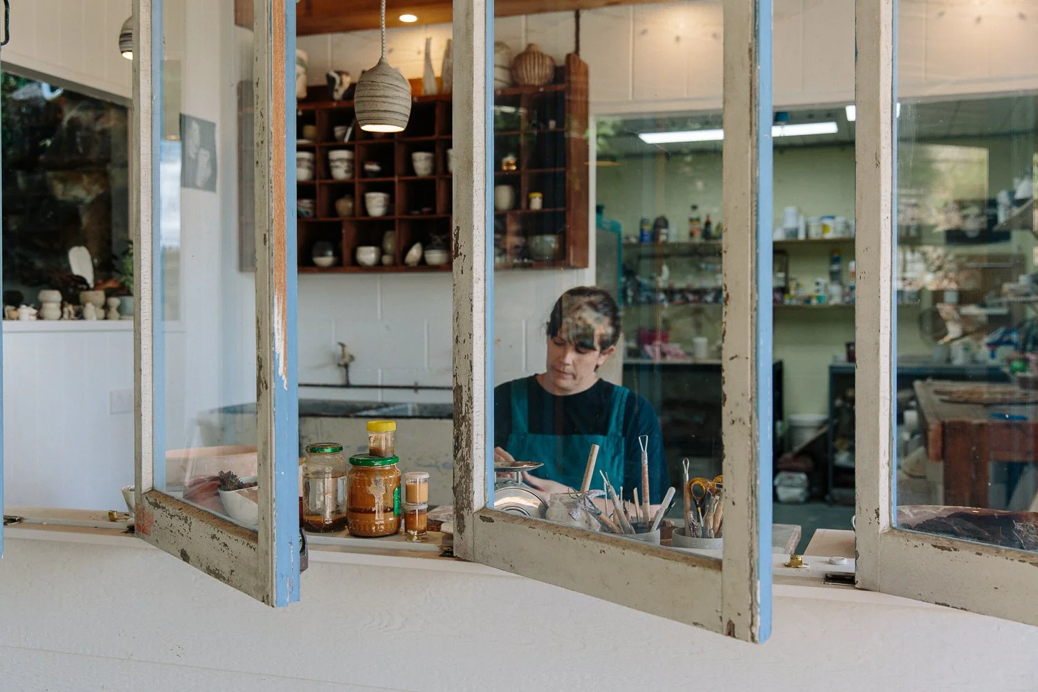 Person working at an artist's or craftsperson's workshop, seen through a window with peeling paint, surrounded by tools, jars, and materials.