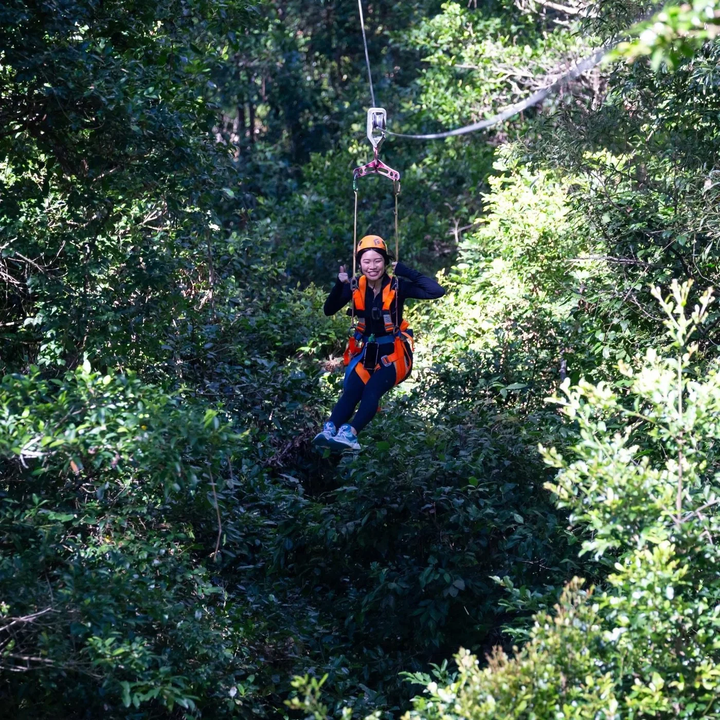 A young woman in an orange helmet and harness zip-lining through a lush green forest.