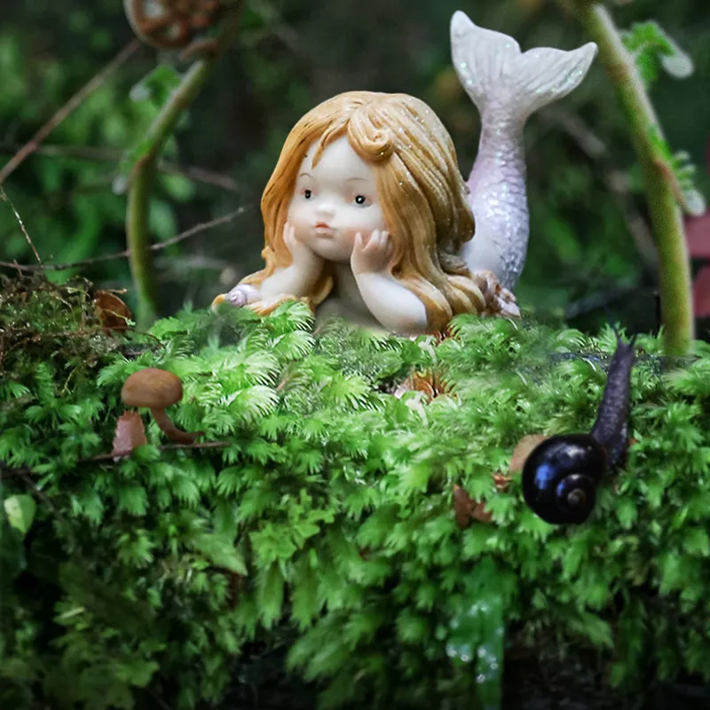 A decorative figurine of a girl with long blonde hair and a mermaid tail, resting on green moss with small mushrooms and a snail nearby, in a garden setting.